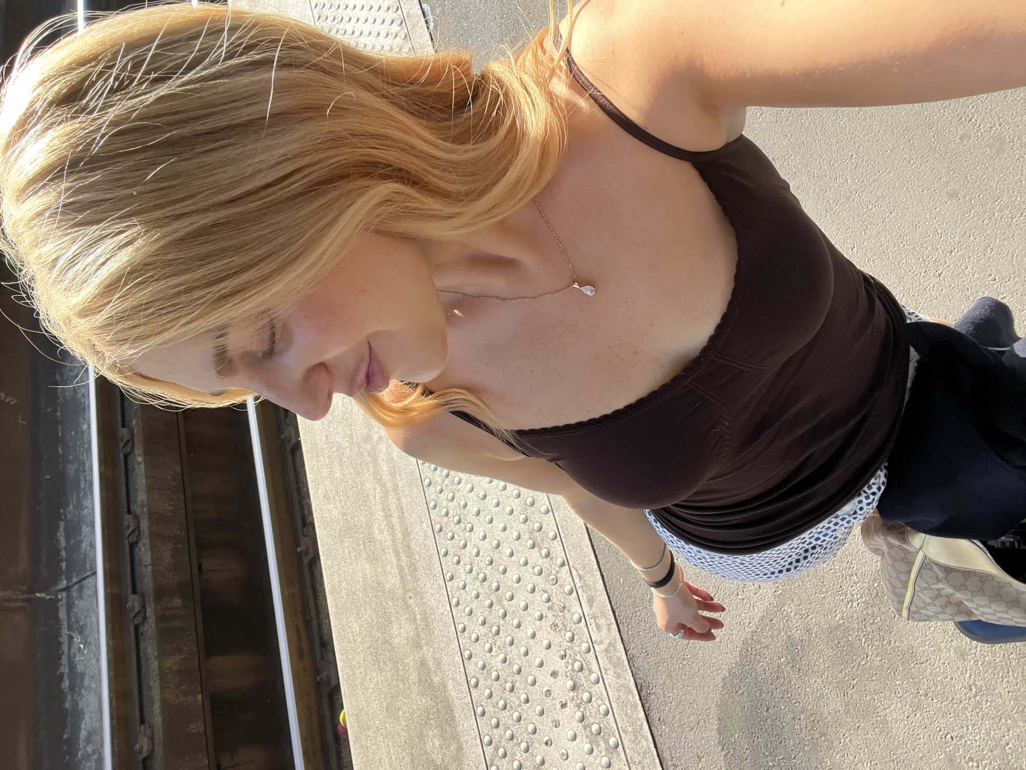 A woman stands on a textured sidewalk near subway tracks, likely in Paris, France, as indicated by the typical style of the subway platform and the style of the subway tracks. She is wearing a brown camisole and a white polka-dotted skirt with dark leggings underneath. She also wears a gold necklace and has multiple bracelets on her left wrist. Her hair is blonde and shoulder-length, styled in waves. A patterned shoulder bag is visible on the ground to the right of the image. The sun is shining brightly, casting shadows on the scene.