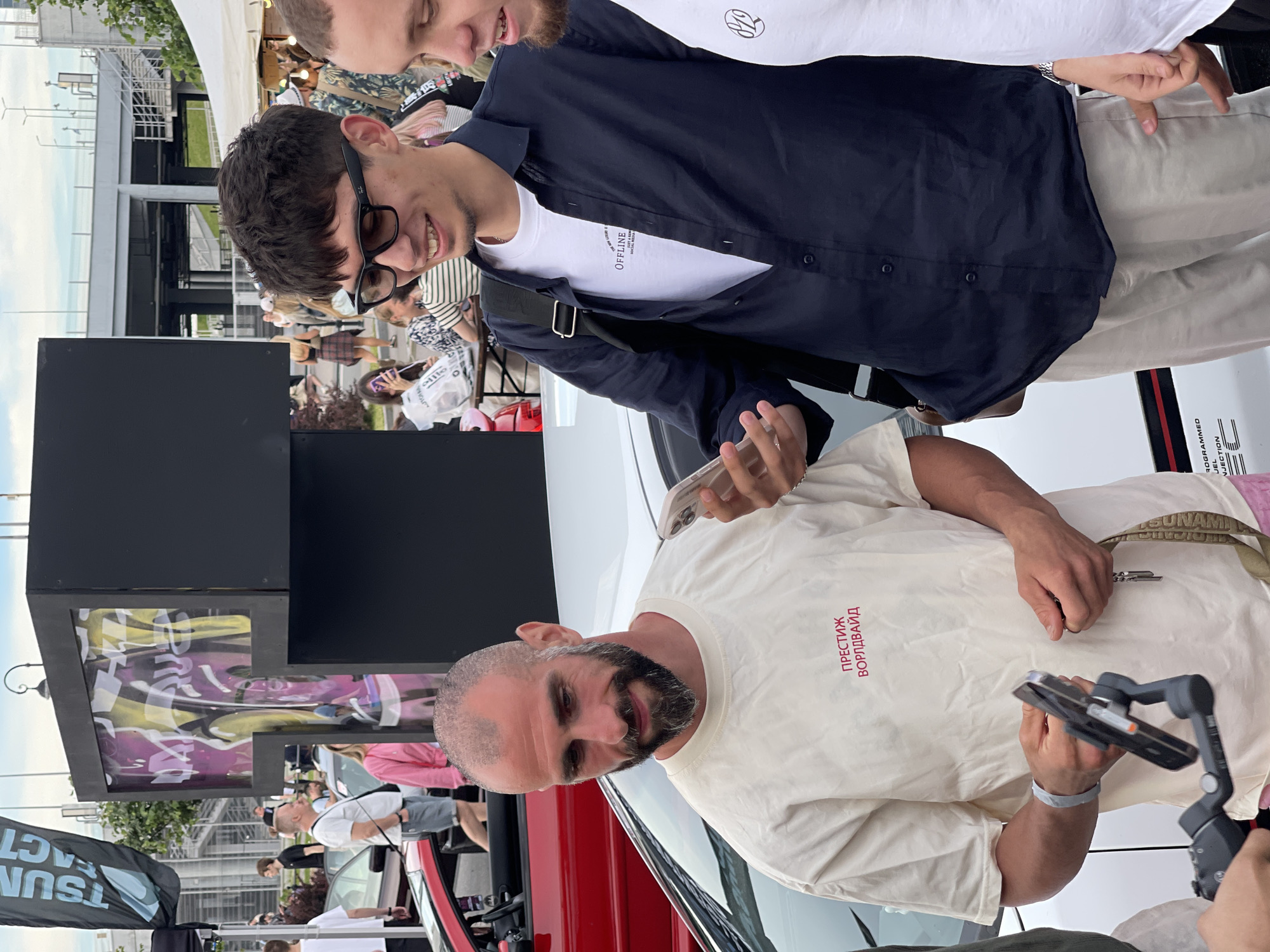 Three men are standing in front of a white car at an outdoor event. The man on the left, with a shaved head and a beard, is wearing a cream-colored t-shirt with red text. He is holding a mobile phone attached to a gimbal. The man in the middle is wearing a dark blue button-up shirt over a white t-shirt and light-colored pants. He is looking at his phone. The man on the right is wearing a white t-shirt and is partially visible. In the background, there is a black structure with graffiti, tents, and other people. The sky is overcast.
