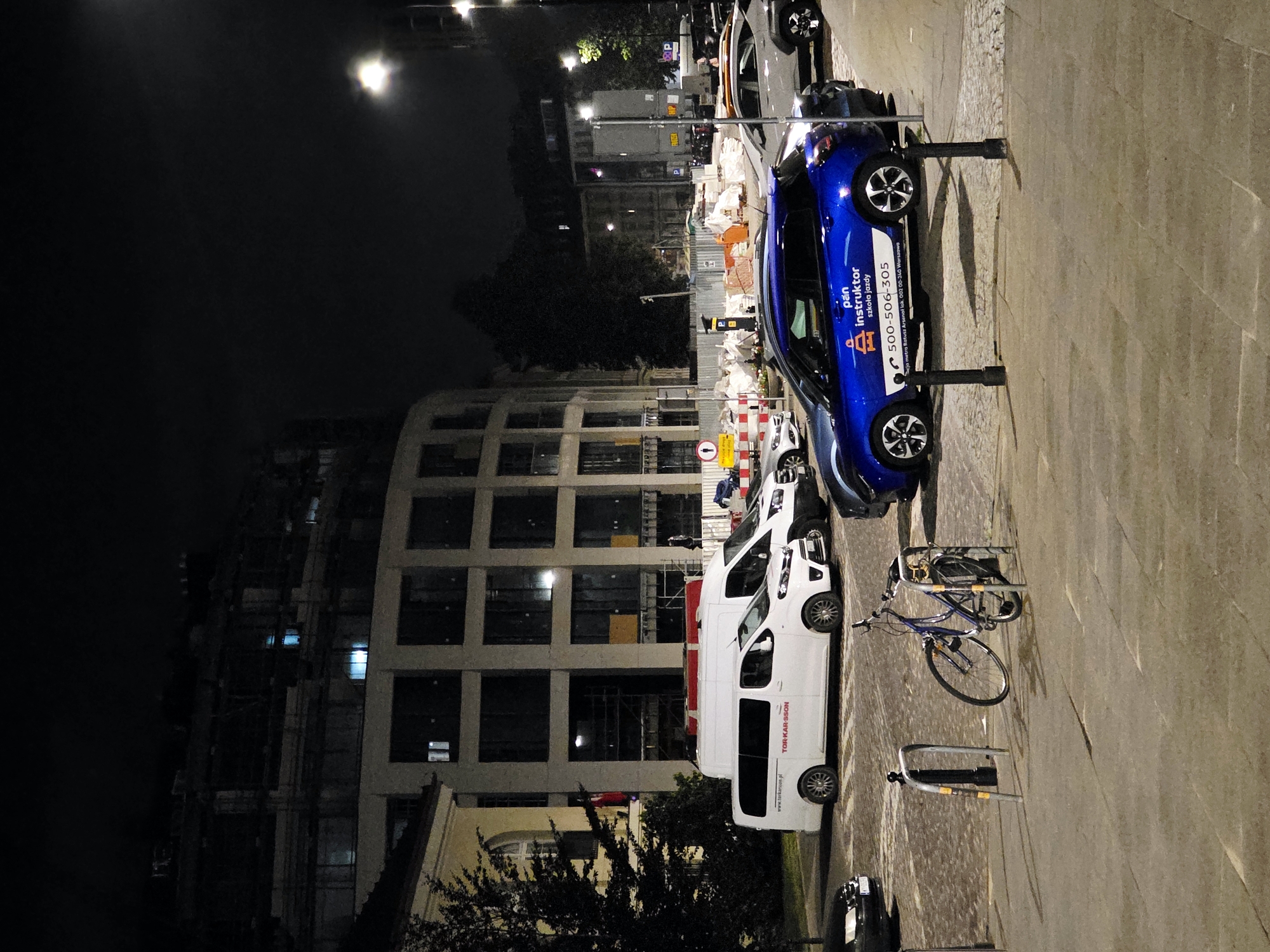 In Warsaw, Poland, at night, a street scene features a partially constructed building in the background with scaffolding visible. In the mid-ground, several vehicles are parked, including a white van with the text "TOR-KAR-SSON" and "www.torkarsson.pl" on its side, another white van, a blue car with "pan instruktor szkoła jazdy" and a phone number printed on it, and several other cars. A bicycle is parked at a bike rack in the foreground. The ground is paved with cobblestones. Streetlights illuminate the scene.