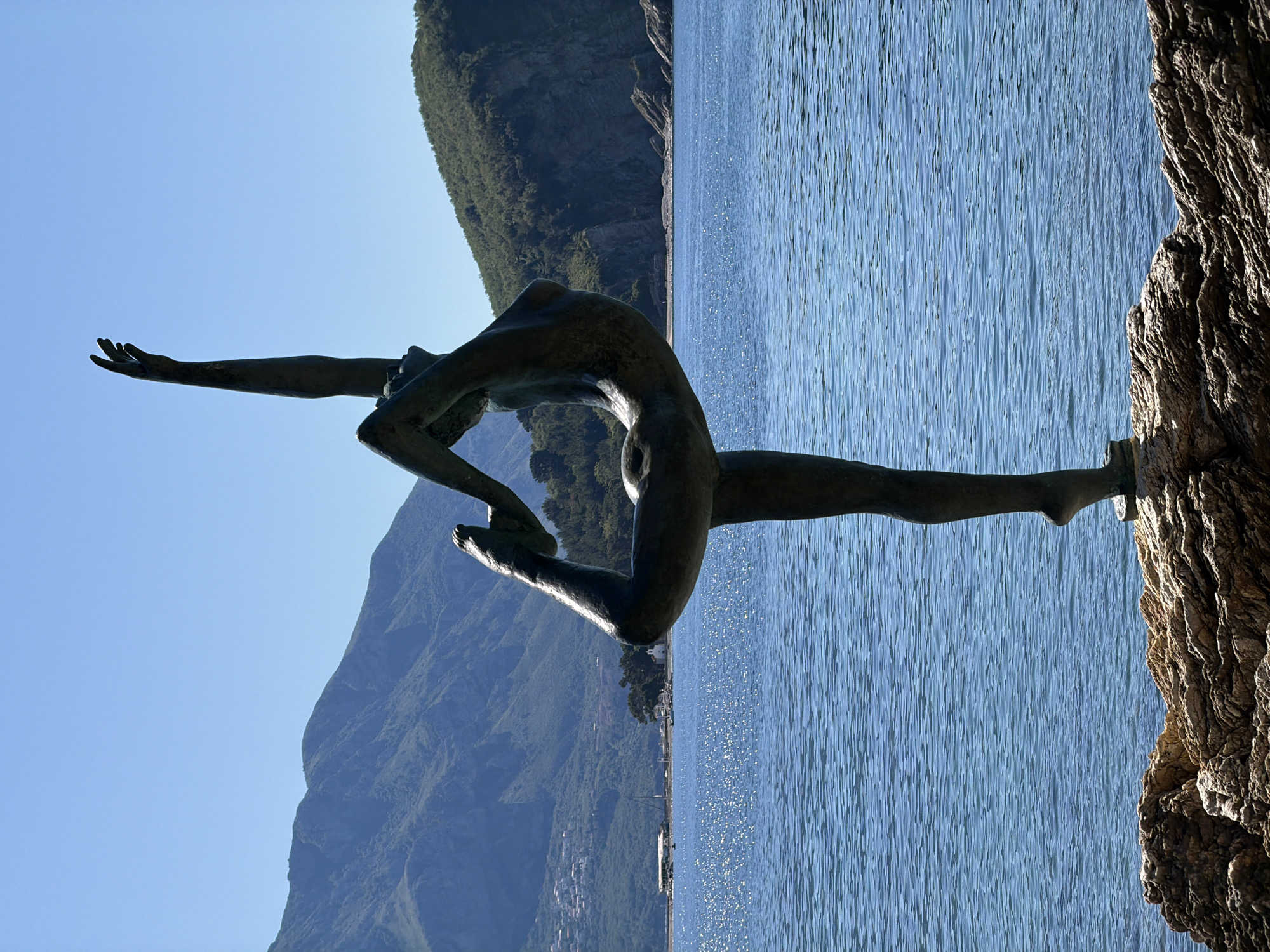 The image shows the "Ballerina of Budva" statue in Montenegro. The bronze statue of a female dancer is perched on a rocky outcrop along the coastline. The Adriatic Sea stretches behind the statue, with mountains visible in the distance. The sky is clear and blue.