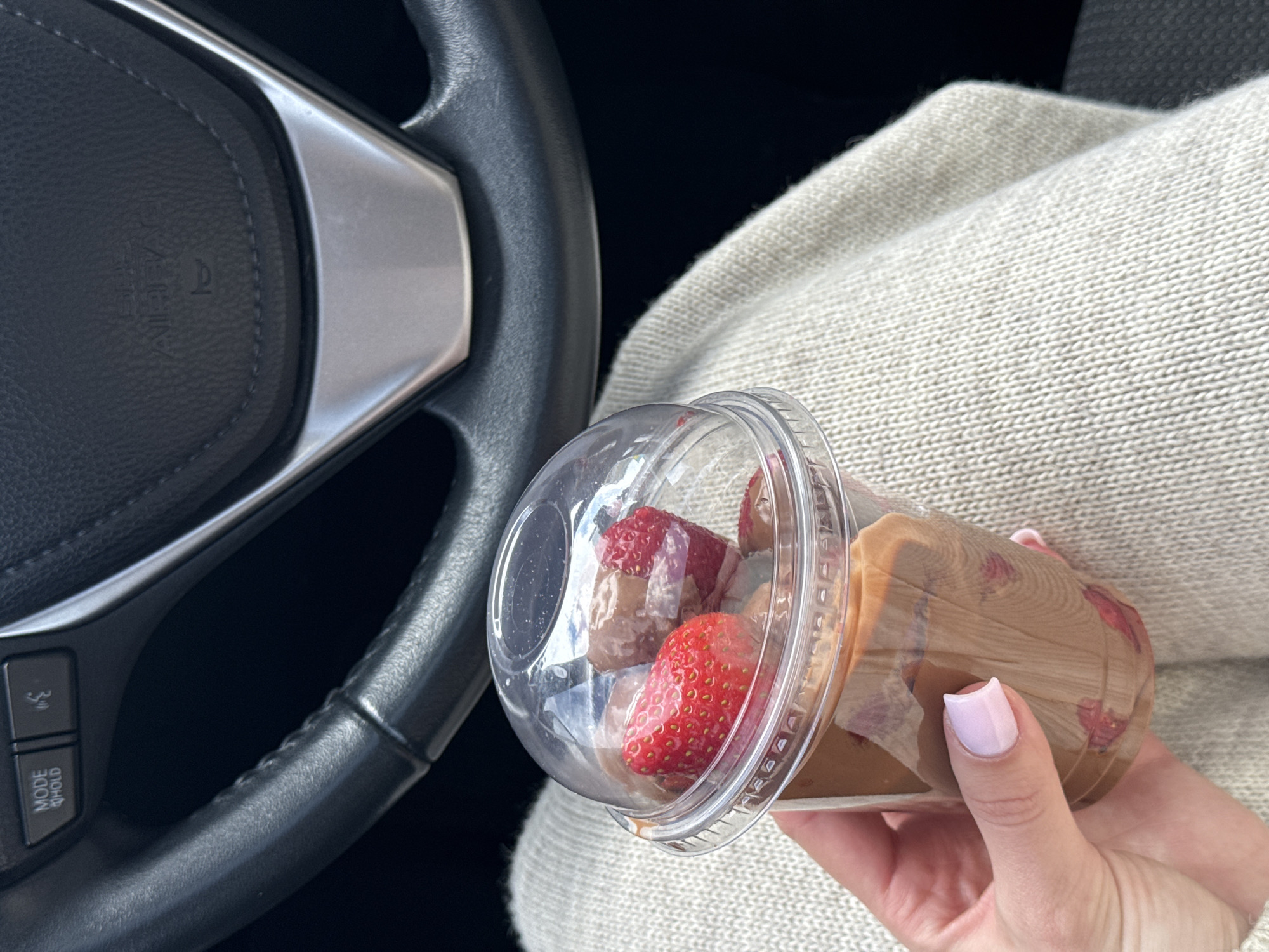 A person holds a plastic cup filled with a brown liquid and strawberries in their hand while inside a car. The steering wheel of the car is visible, as is the person's light-colored sweater. The image does not provide any information to identify the location as Kyiv, Ukraine.