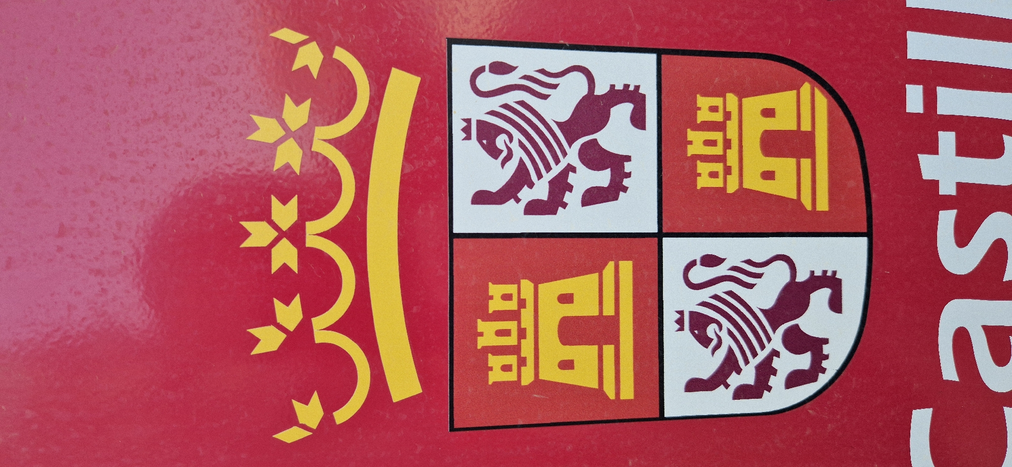 The image displays a close-up of a red surface, likely a sign or banner, featuring a coat of arms. The coat of arms is divided into four quadrants, topped by a yellow crown with red accents. The upper left and lower right quadrants depict yellow castles with red windows and doorways. The upper right and lower left quadrants show maroon lions with red manes and striped bodies. Below the coat of arms, the word "Castil" is visible in large white serif letters. The background is a solid red with a subtle speckled texture, suggesting a painted or enameled surface. The lighting is even, with no harsh shadows, indicating a likely indoor setting or overcast conditions. No people are visible in the frame.