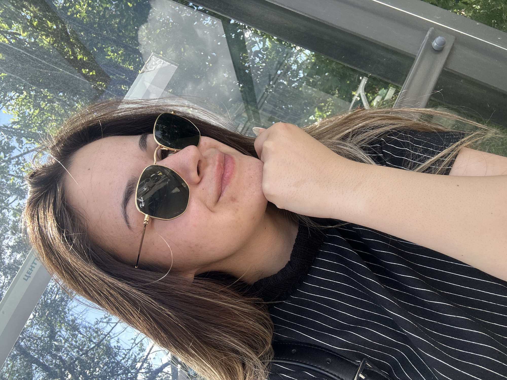 A young woman poses for a selfie. She wears sunglasses with a gold frame, a black and white striped shirt, and sits in an enclosed glass structure. The glass appears to be the wall of a bus stop, as implied by the structure and trees reflected in the glass. The location is in Sofia, Bulgaria.