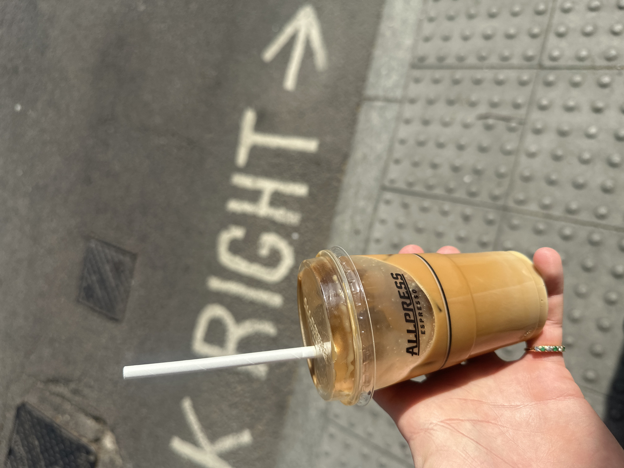 A hand holds an Allpress Espresso iced coffee on a sidewalk. The sidewalk is grey concrete with a directional arrow and the word "RIGHT" painted in white. The sidewalk transitions to a textured, nubbed surface typical of pedestrian walkways in London.