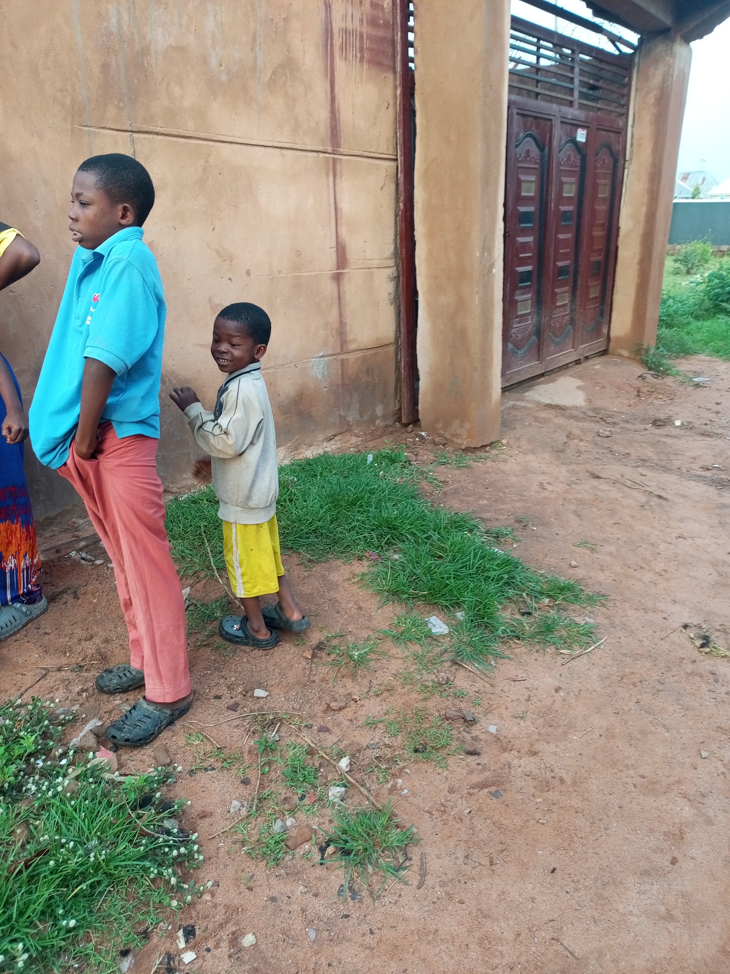 Two young boys stand on a dirt path in front of a weathered, tan building with a dark red metal gate. The boy on the left, wearing a bright blue polo shirt and salmon-colored pants, looks to his left with his hands in his pockets. The boy on the right, wearing a light grey long-sleeved shirt and yellow shorts, is facing the viewer with a wide smile and is captured in mid-motion, possibly talking or gesturing. He wears dark clogs. The ground is covered in dirt with patches of green grass and scattered debris. The building's facade appears rough and textured, with horizontal lines etched into it. The gate, composed of multiple vertical panels with decorative elements, is closed. In the background, beyond the gate, a grey fence and some greenery are visible. The scene suggests an outdoor setting, possibly a residential area or a communal space, in Tempure, Nigeria.