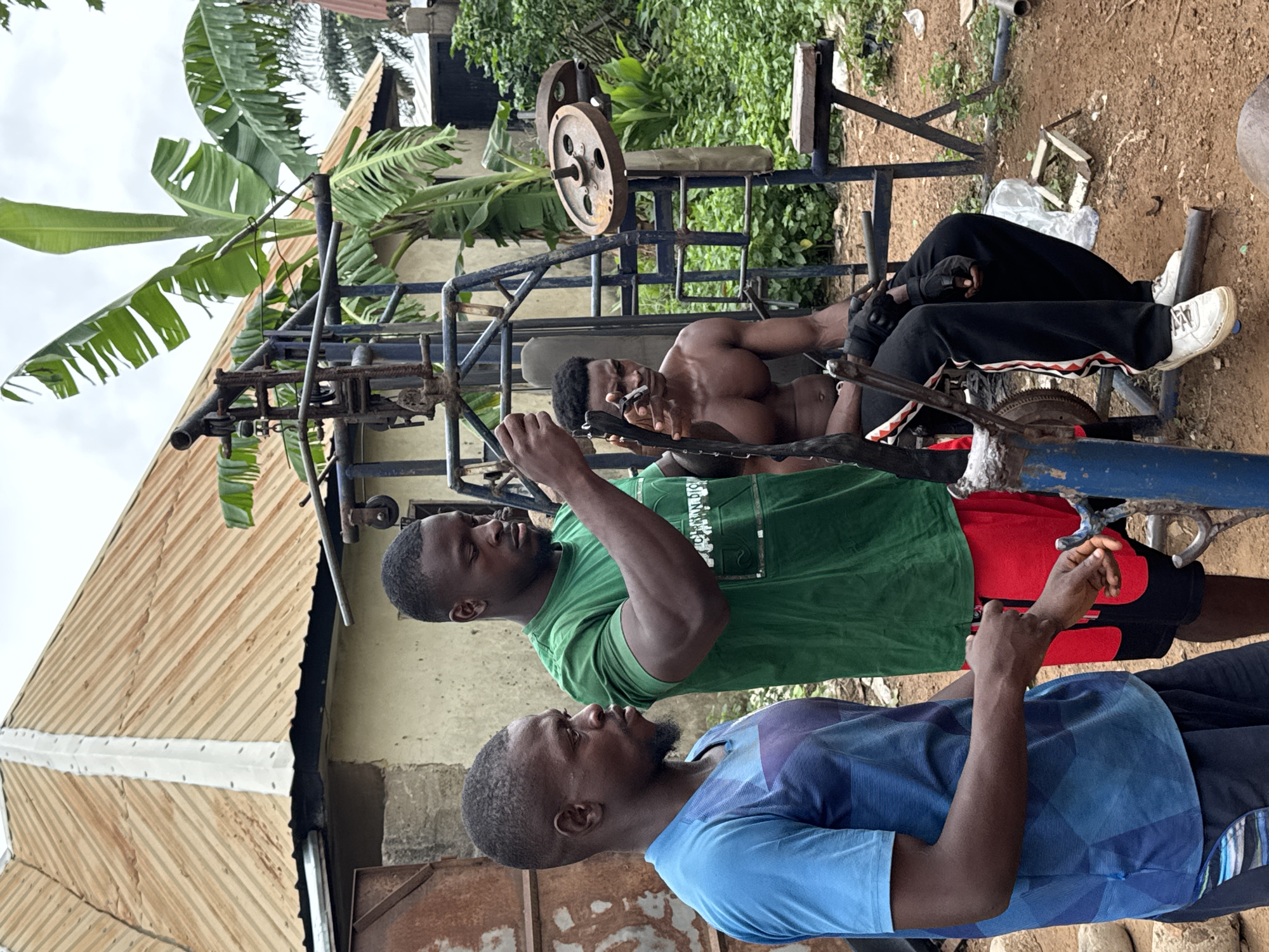 In Akwanga, Nigeria, three men are present in an outdoor setting. Two men are standing, and one is seated. The man on the left, wearing a blue t-shirt and dark shorts, has short hair and a beard. The man in the center, wearing a green t-shirt and red shorts, is muscular with short hair. He is interacting with the seated man, who is shirtless and wearing black track pants and white sneakers. The seated man has a muscular build and appears to be using a weightlifting apparatus. The weightlifting equipment is made of metal and has visible weights. The background shows a building with a corrugated metal roof, banana trees, and lush green vegetation. The ground is covered in reddish-brown dirt. The scene appears to be a makeshift outdoor gym.
