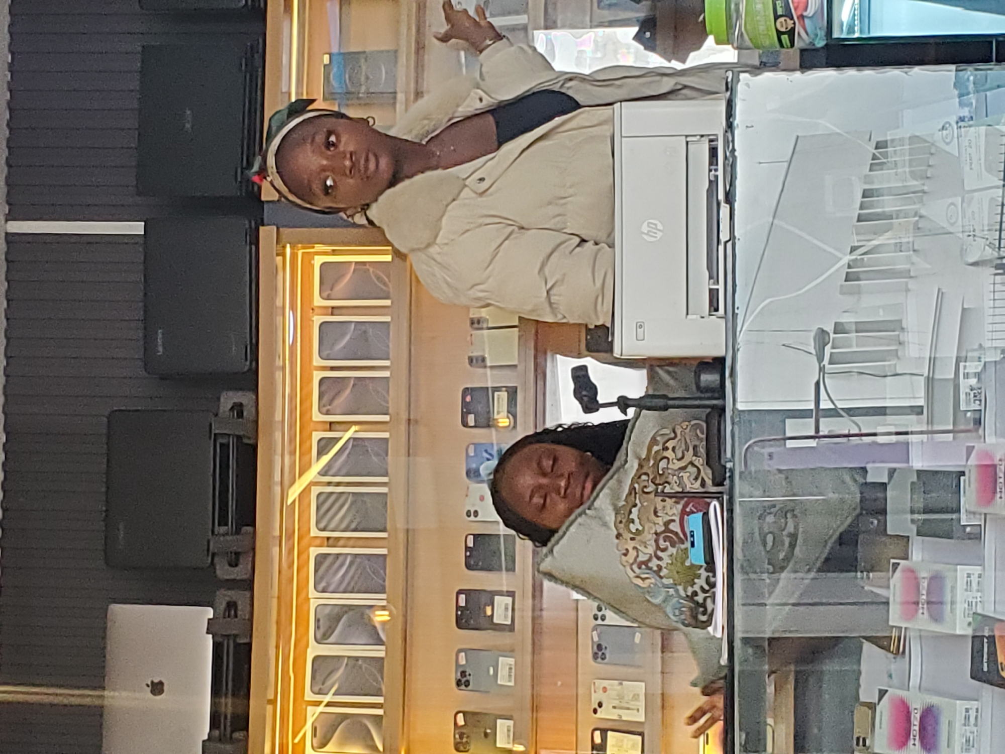 Two young Black women are the subjects of this photo, which appears to be taken inside an electronics store. The woman on the left is standing, wearing a light-colored puffer jacket over a dark top and a green and white headscarf. She is looking directly at the camera with a surprised expression, her left hand raised slightly. Behind her, a white HP printer is visible.
The woman on the right is lying down, her head resting on a decorative cushion with an ornate pattern. She is wearing a gray jacket. Her eyes are closed, suggesting she is sleeping or resting.
The setting is a store displaying numerous cell phones, likely new ones in boxes, arranged in vertical rows on shelves. The shelves are illuminated by warm lighting, creating a reflective effect on the glass front of the display. In the background, dark, flat objects, possibly laptop boxes, are stacked on shelves. The overall lighting suggests it could be daytime, as the store is brightly lit. The reflections on the glass indicate the photo is taken from outside the display, possibly through a window or glass partition. The presence of cell phones and electronics suggests the location is an electronics retail store. The location context is Jos, Nigeria. Visible text includes "HP" on the printer and some unreadable text on product packaging.