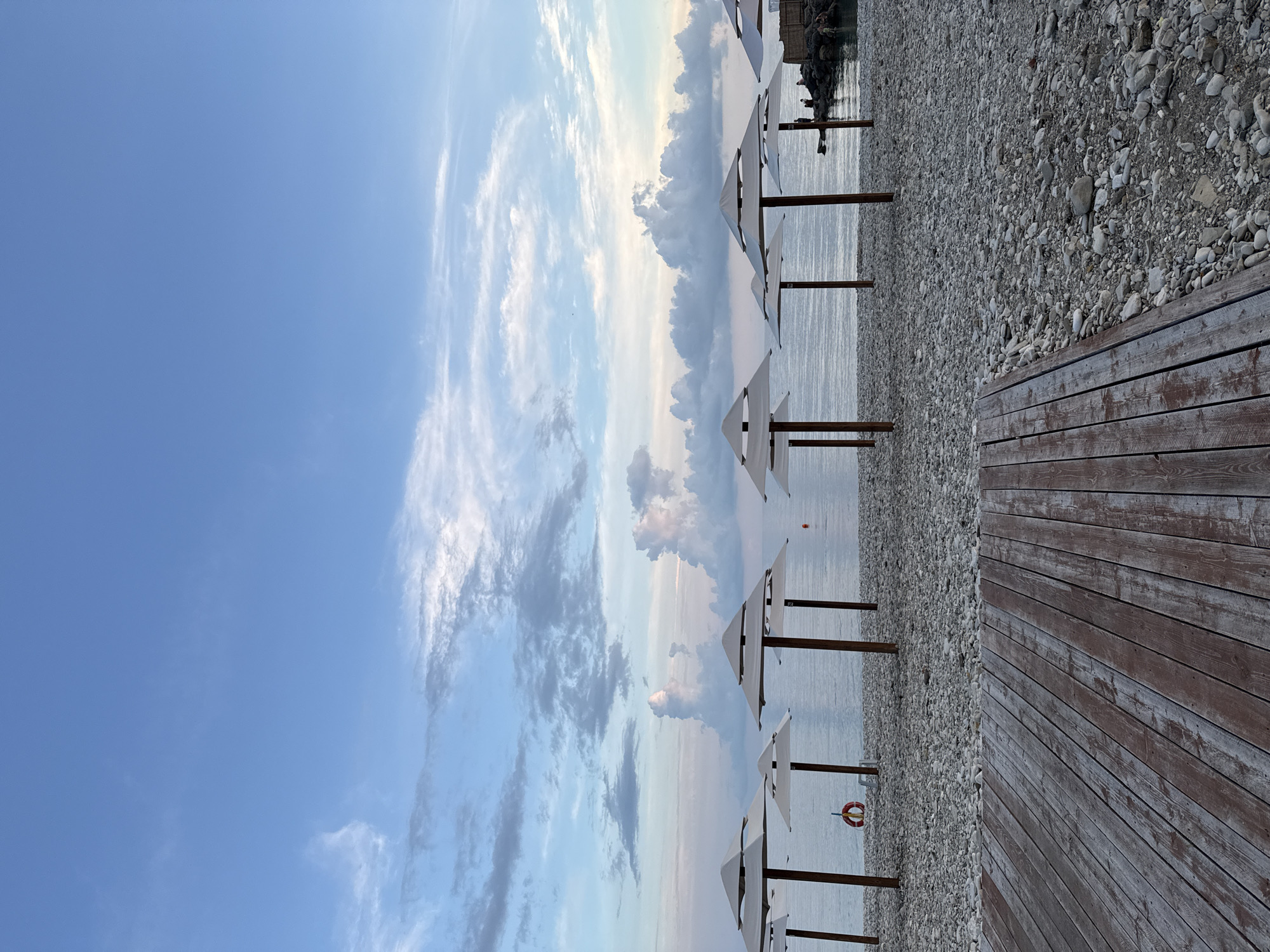 The image depicts a serene beach scene on a pebbly shore in Nebug, Russia, at what appears to be late afternoon or early evening. A weathered wooden boardwalk occupies the foreground, leading towards the sea. Rows of white, modern beach umbrellas are positioned on the pebble beach, with their wooden poles firmly planted in the ground. The sea is calm and reflects the soft hues of the sky, which is a gradient of pale blue with scattered, wispy clouds tinged with pink and orange. In the middle ground, a red life preserver hangs on a pole. Further to the right, a rocky breakwater extends into the water, with a few indistinct figures standing on it. The overall atmosphere is peaceful and tranquil, suggesting a quiet moment before sunset.