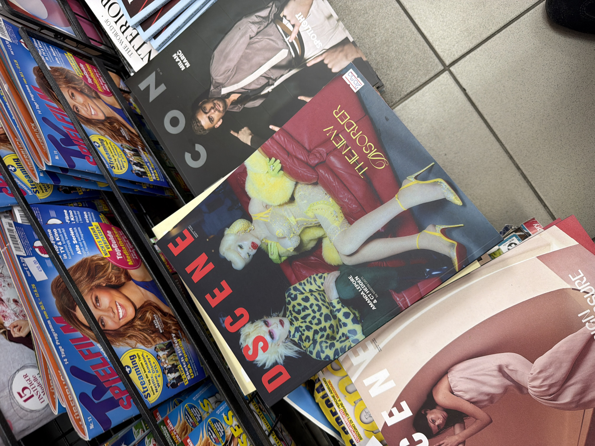 The image shows a display of magazines in a rack, likely at a newsstand or bookstore. Several magazine covers are visible, including "Spiel Film", "Cono", "Dscene" and "Design Cure". The "Dscene" magazine cover features a photograph of Amanda Lepore & CT Hedden. The location cannot be identified as Belgrade, Serbia from the image. The environment appears to be indoors, possibly a retail space.