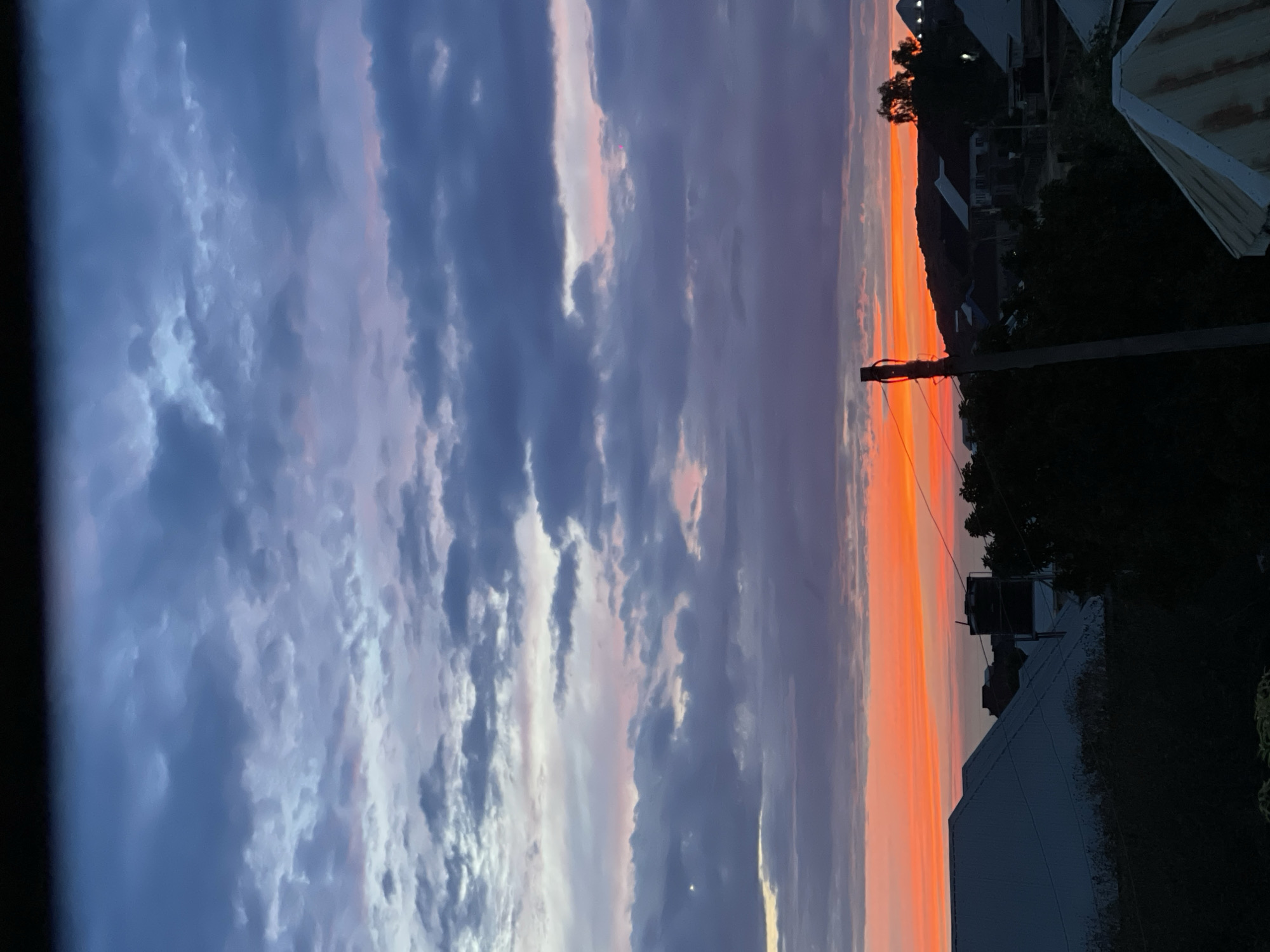 The image depicts a sunset with orange and purple hues, likely in the city of Jos, Nigeria, based on surrounding structures. The sky is filled with clouds. A utility pole and power lines are visible against the setting sun. Houses and trees are silhouetted in the foreground. No people are visible.