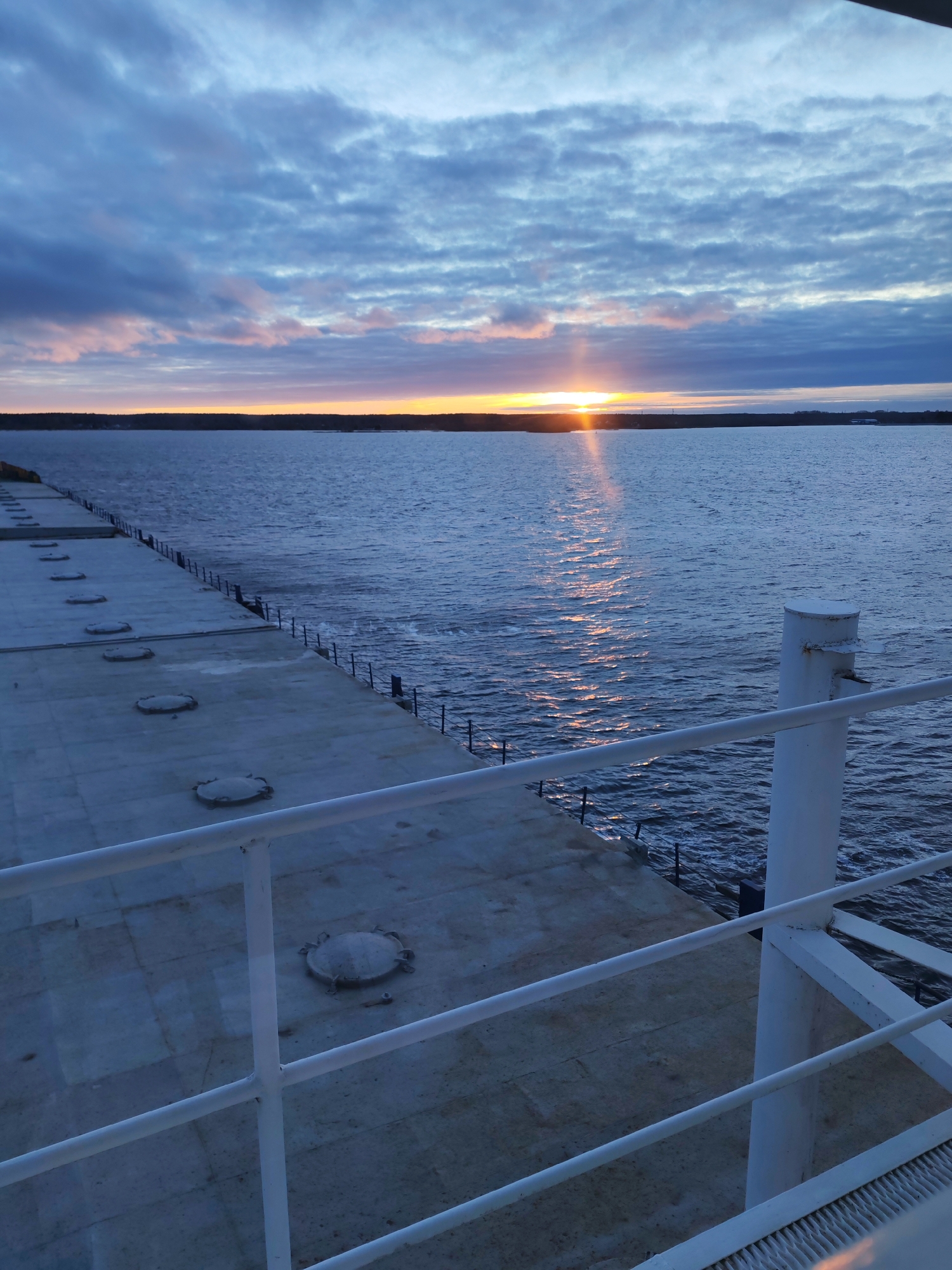The image presents a serene view of a sunrise or sunset over a large body of water, possibly a river or lake in the Bol'shie Mkhi area of Russia. The scene is observed from the deck of what appears to be a ship or a large waterside platform.
In the foreground, a sturdy white railing diagonally traverses the lower right portion of the frame, providing a visual anchor. To the left, a broad, flat deck, possibly made of concrete, extends into the distance, lined with a series of regularly spaced, round, raised covers or hatches. Along the edge of this deck, a darker, lower railing or fence separates it from the water.
The water itself is a deep blue, exhibiting gentle ripples and small waves. A brilliant sun, low on the horizon, casts a radiant, golden path of light across the water, creating a shimmering reflection that draws the eye towards the vanishing point.
The horizon features a strip of distant land, likely covered with dense forest, silhouetted against the glowing sky. Above, the sky is dominated by a scattering of stratocumulus clouds, colored in shades of blue and grey, with soft pink and orange hues near the sun, characteristic of either dawn or dusk. The overall impression is one of peaceful natural beauty. No human activity or visible text is present in the image.