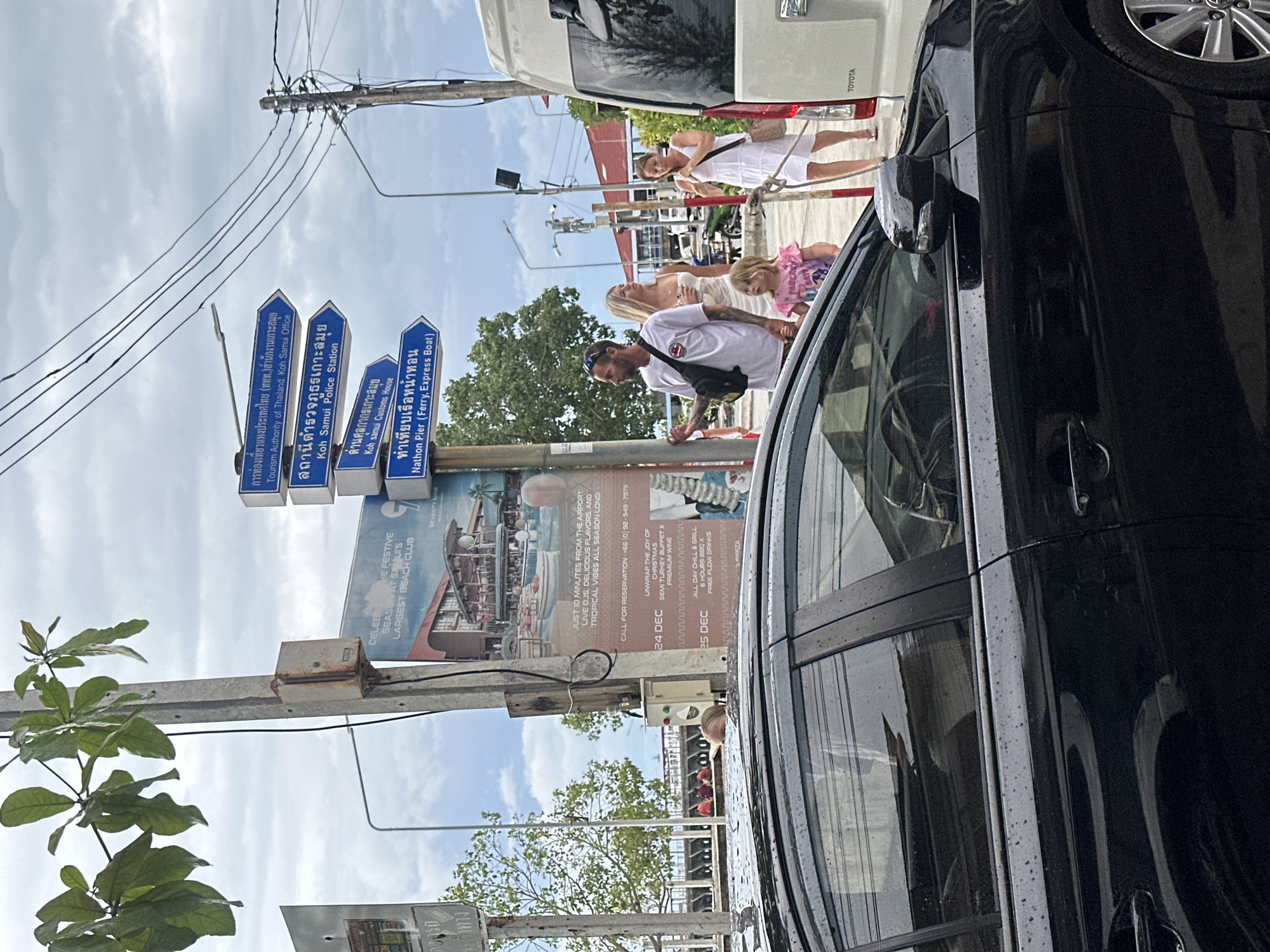 This image captures a street scene on Koh Samui, Thailand, near Nathon Pier, as indicated by visible signage, aligning with a location such as Baan Lipa Noi. The foreground is dominated by the side profile of a black sedan, possibly a Toyota, which appears wet with small droplets, suggesting recent rain. The driver's side front and rear doors, window frames, and part of the front wheel and side mirror are visible.
In the midground, a group of four Caucasians stands on a paved surface, likely a sidewalk. A man with a beard and sunglasses pushed up on his head wears a white t-shirt and a black fanny pack; he holds the hand of a small child, a girl in a pink t-shirt. Standing next to them is a woman with blonde hair in a white strapless dress. To their right, another woman in a white dress carries a light-colored handbag and looks towards the left. Behind this group, the rear end of a white Toyota passenger van is partially visible.
Further into the midground, a concrete utility pole displays four blue directional signs with text in both Thai and English. From top to bottom, they point to the "Tourism Authority of Thailand, Koh Samui Office," "Koh Samui Police Station," "Koh Samui Customs House," and "Nathon Pier (Ferry, Express Boat)." Attached to the pole is a rusty electrical box with exposed wiring. A large billboard poster is partially visible behind the pole, advertising "SAMUI'S LARGEST BEACH CLUB" with promotional text about festive season events on "24 DEC" and "25 DEC."
The background reveals a paved area leading to a railing, behind which red objects are visible. Buildings with red roofs and overhead power lines are discernible further back. The sky is overcast with scattered clouds. A branch with green leaves from a tree partially obstructs the upper left portion of the frame.