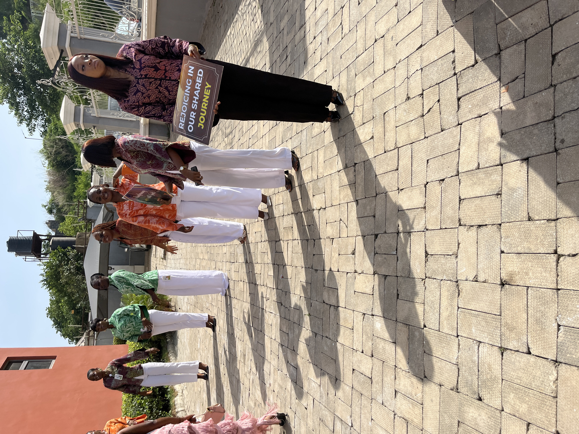 A group of women stand in a line on a paved area outdoors, likely in Abuja, Nigeria, given the context provided. The paving is made of light-colored, rectangular blocks. The women are wearing matching outfits: patterned tops with varying colors, and white pants. They all wear dark-colored footwear. One woman at the front of the line holds a sign that reads "REJOICING IN OUR SHARED JOURNEY". The woman holding the sign wears black pants. The shadows cast by the people suggest a bright, sunny day. In the background, a building with a water tank on top is visible, along with green trees and a fence.