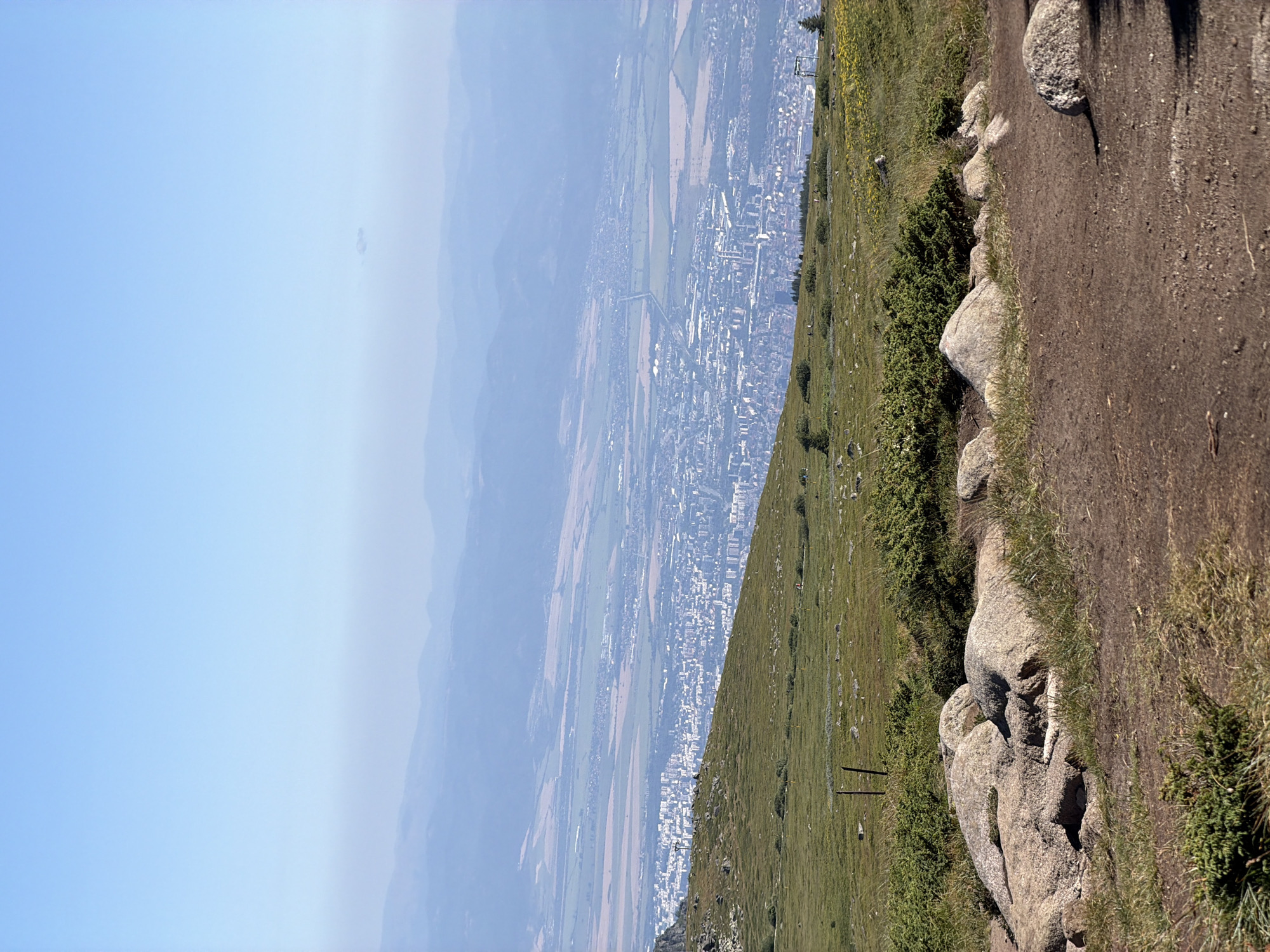 The image shows a panoramic view from a high vantage point, overlooking a sprawling city nestled in a valley. The foreground features a rocky, grassy terrain with scattered boulders and low-lying green shrubs. A dirt path winds through this area. Beyond the foreground, the landscape transitions to a green, hilly expanse dotted with more shrubs and some scattered yellow wildflowers. Further in the distance, the city, which appears to be Bistritsa, Bulgaria, unfolds with a dense concentration of buildings. The city is situated within a vast plain, bordered by several layers of hazy, blue-toned mountains receding into the horizon under a clear, pale blue sky. The overall impression is one of a vast, scenic landscape with human settlement integrated into the natural environment.