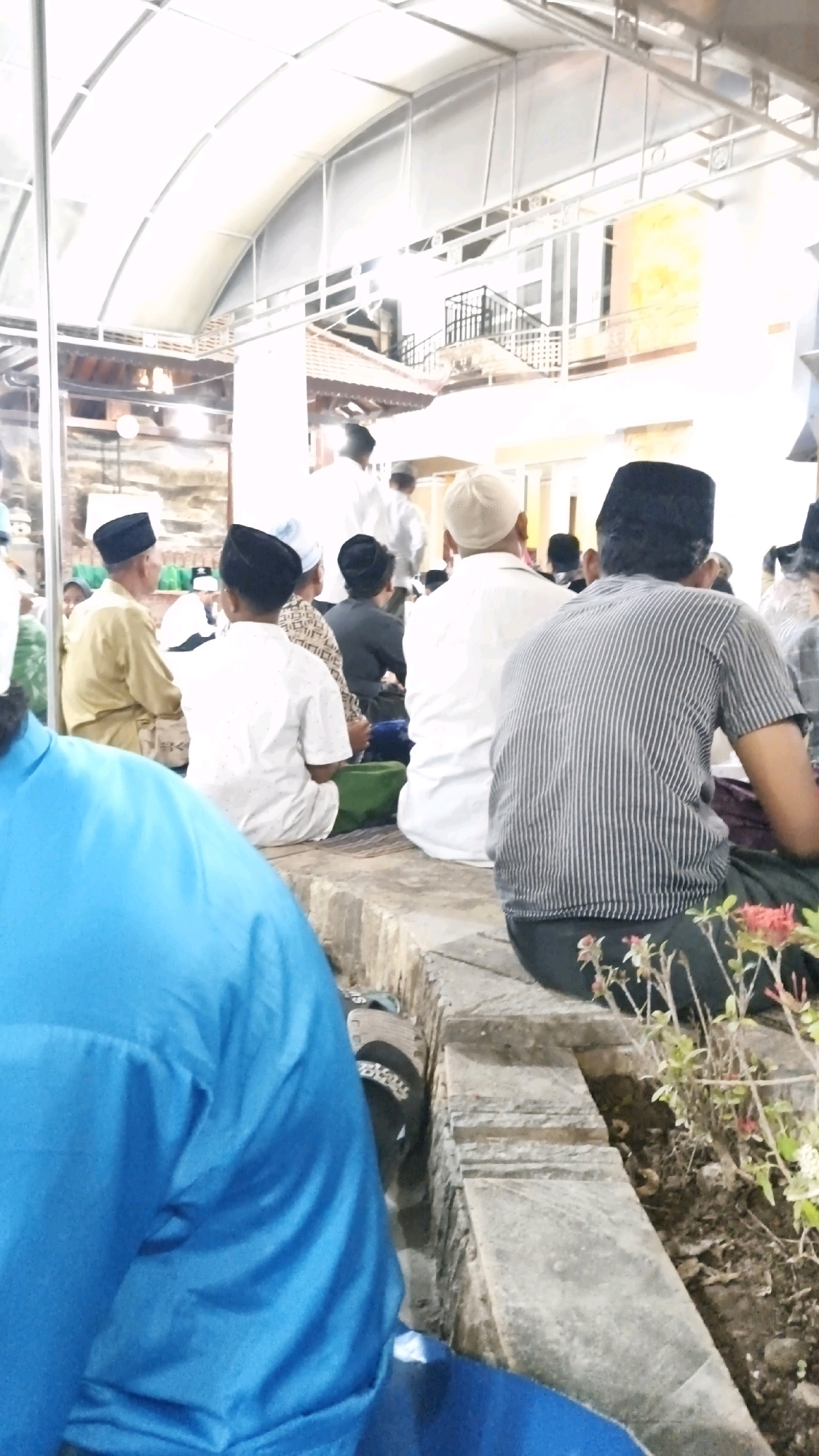 This image depicts an outdoor gathering of people, likely a religious event, taking place in Sambipondok, Indonesia. The individuals are seated on a stone ledge, facing away from the camera in most cases, suggesting they are observing something or engaged in a shared activity.
The environment appears to be a partially covered area, with a large, white, translucent roof structure overhead, supported by metal beams. Below the roof, traditional Indonesian architectural elements are visible, including tiled roofs and wooden structures. The ground is uneven, with a mix of stone and dirt. Small plants with red and white flowers are growing near the stone ledge in the foreground.
The people in the image are dressed in various attire, with many wearing traditional Islamic clothing, including white shirts, peci (caps), and sarongs. One individual in the foreground is wearing a bright blue shirt. The overall mood of the scene is calm and communal. The lighting suggests it is daytime, with natural light filtering through the roof.