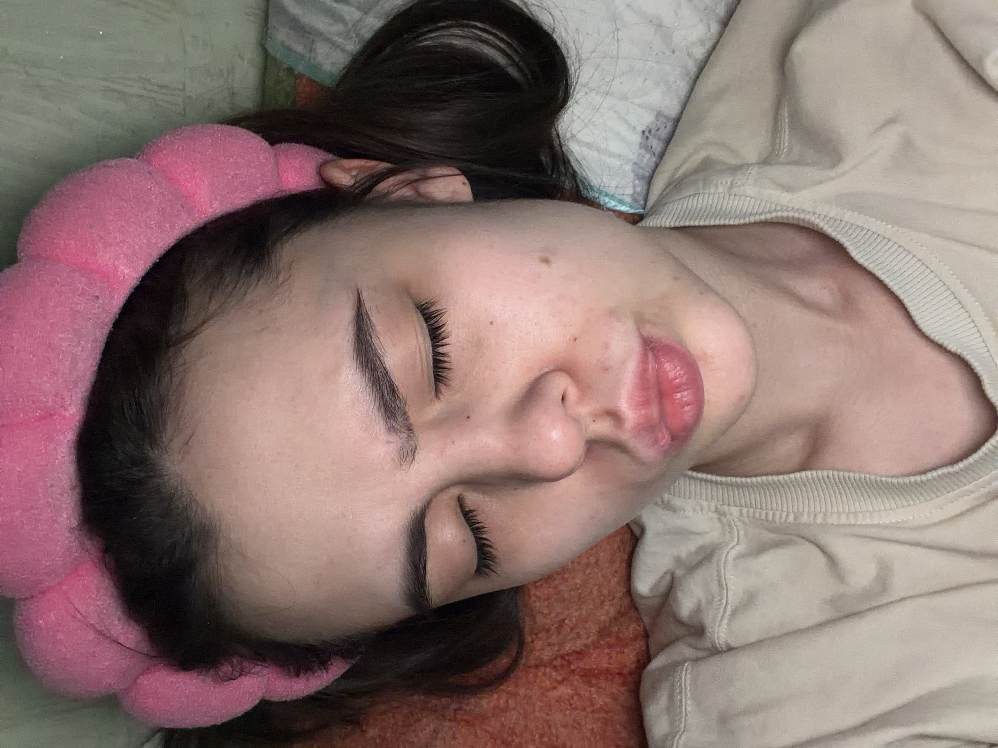 A woman is lying down, presumably sleeping, with her head turned to the left. She has dark hair, fair skin, and is wearing a pink, puffy headband. She is wearing a light-colored, long-sleeved shirt with a ribbed collar. She is lying on what appears to be a bed or couch with a patterned pillow and reddish-brown fabric underneath. There is no information available in the image that identifies the location as the city of Chaikovsky, Russia.