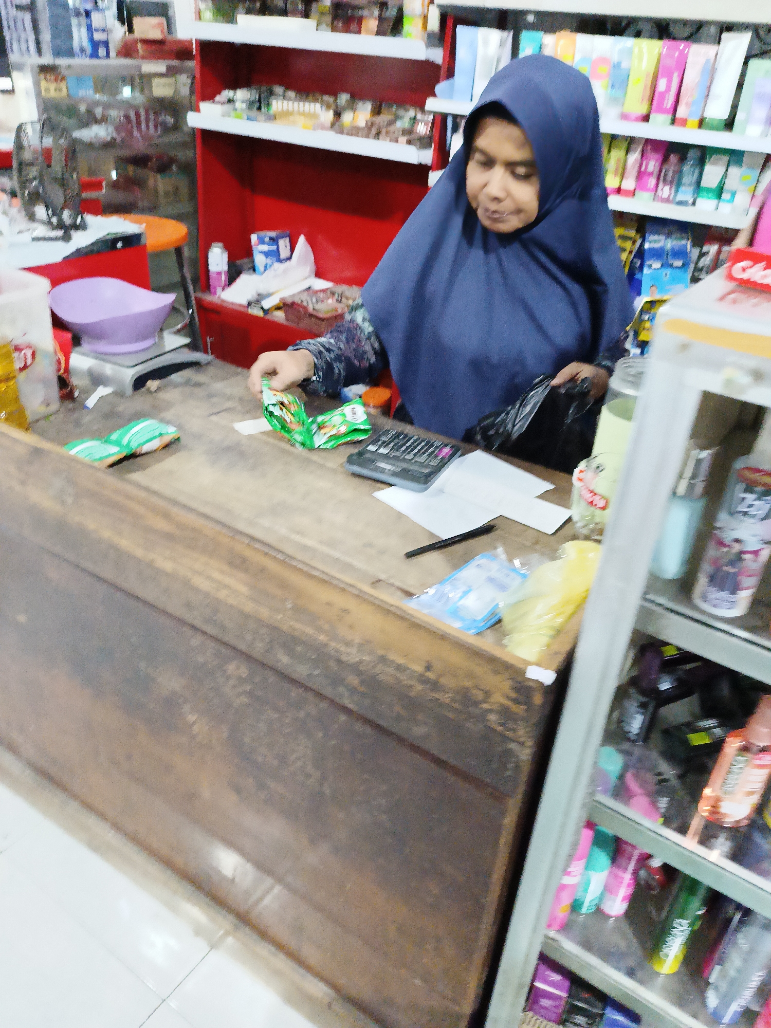 A woman wearing a dark blue hijab is standing behind a wooden counter in a small store. She is looking down and appears to be handling a small, green, foil packet of snacks. To her right, a calculator and some white papers are visible on the counter. Behind her, shelves are stocked with various products, including toiletries and food items. The store appears to be a local convenience store, possibly in Sukorini, Indonesia. The lighting is bright, typical of an indoor retail space.