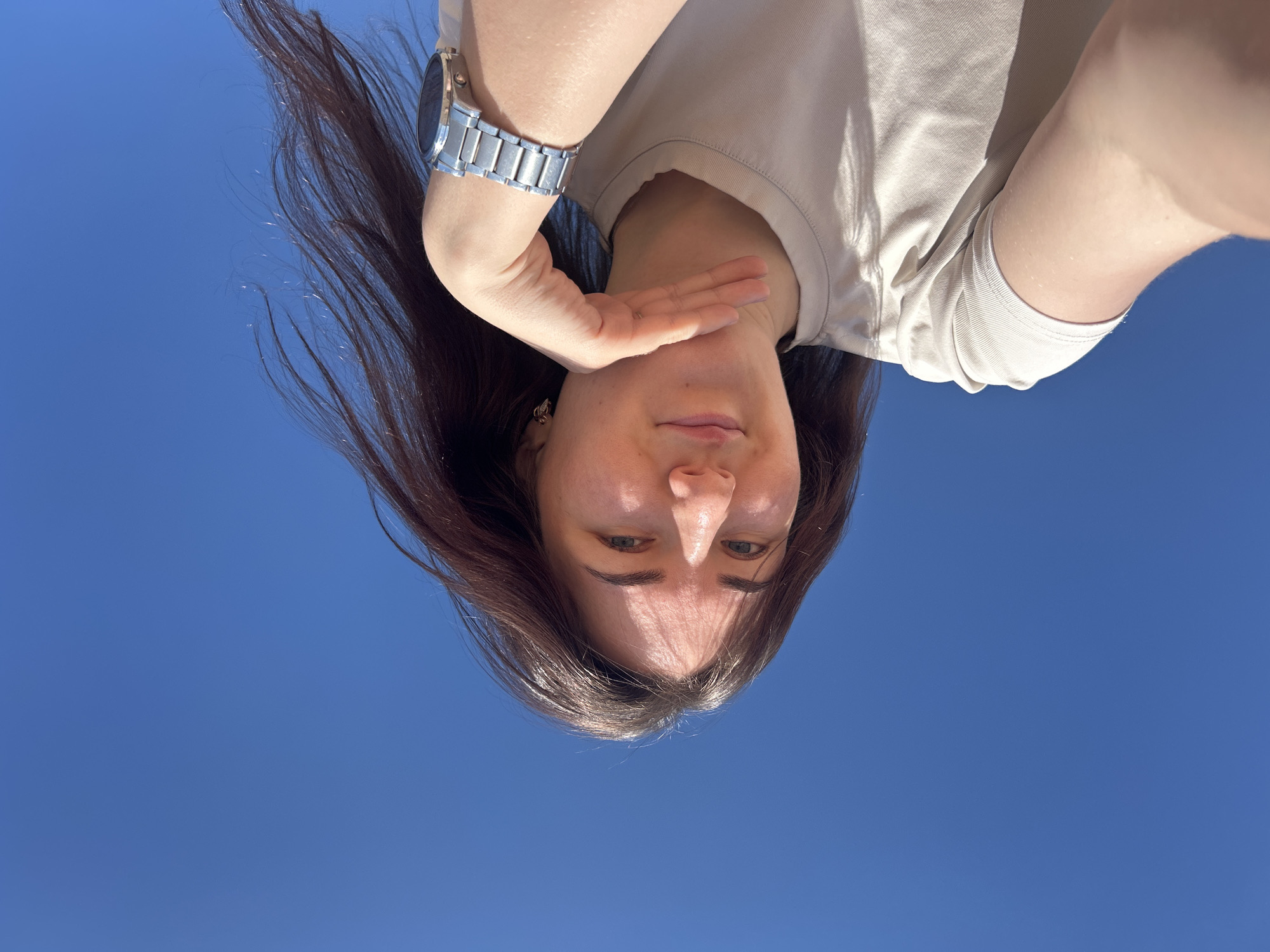 A young woman is photographed from a low angle against a clear blue sky. Her long brown hair is partially windblown, and she is wearing a beige t-shirt. She has a metal wristwatch on her left wrist and her right hand is near her face. The location cannot be determined as Noviy, Russia from the image alone.