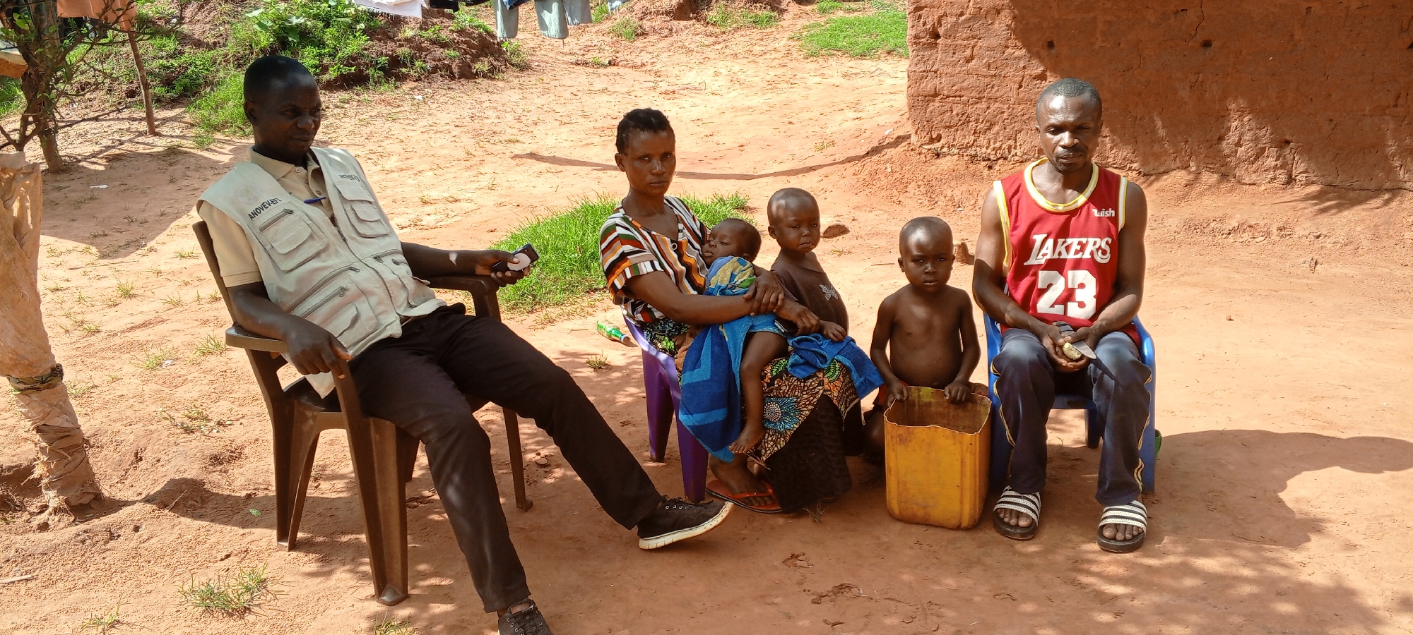 In Ngalula, Democratic Republic of the Congo, a group of five people are gathered outdoors. A man in a khaki vest is seated on a brown plastic chair to the left, holding what appears to be a mobile device. A woman sits next to him on a blue plastic chair, holding a young child wrapped in a blue towel. Beside her sits another child with a dark brown shirt. To the right, a shirtless child sits on a yellow plastic container. Another man, wearing a red Los Angeles Lakers jersey with the number 23, sits on a blue plastic chair to the right of the shirtless child. All are in a yard with sandy soil and sparse grass. A mud brick building is visible in the background to the right.