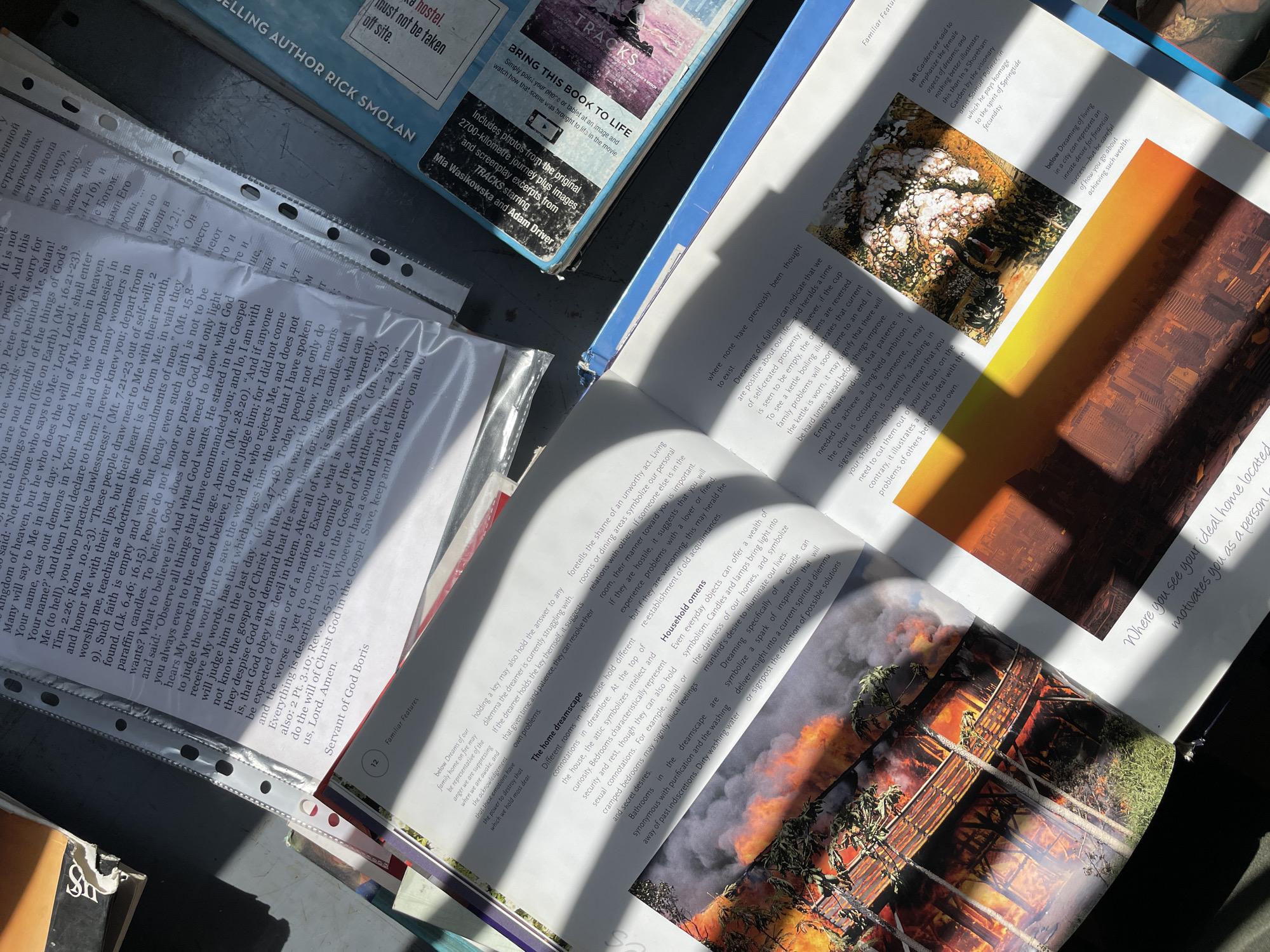An overhead shot captures a collection of books and papers on a dark surface, bathed in bright, directional sunlight creating strong shadows. The scene implies an indoor setting, likely a desk or table, in Tbilisi, Georgia, during a sunny day.
On the far left, a book with a dark cover displays text including "SELLING AUTHOR RICK SMOLAN," "hostel," and "must not be taken off site." Beside it, white papers held within a transparent binder contain extensive religious text, specifically Christian scripture with numerous Bible verse references (e.g., Matthew 16:21-23, John 12:47-48, Revelation 9:15-19), discussing topics like faith, judgment, and the Gospel. The phrase "Servant of God Boris" is visible, along with some Cyrillic script.
Dominating the right side of the image is an open, illustrated book titled "Familiar Features." The left page includes sections on "The home dreamscape" and "Household omens," discussing dream interpretations related to rooms (attic, bedrooms, bathrooms) and everyday objects (cups, kettles, chairs, candles, lamps). The right page features two images: one depicting a white, cloud-like floral arrangement against a dark background, and another showing an industrial or city landscape under a dramatic orange sky. Further down, a vibrant image shows a large structure engulfed in flames and smoke, with green foliage in the foreground. Text alongside these images mentions artists like Jeff Gardens and Samuel Palmer, and discusses interpretations of dreams about living in a city or the ideal home. Another visible book, partially obscured, is titled "TRACKS" and advertises features like "BRING THIS BOOK TO LIFE" with interactive content, and "Includes photos from the original 2700-kilometre journey plus images and screenplay excerpts from TRACKS starring Mia Wasikowska and Adam Driver."