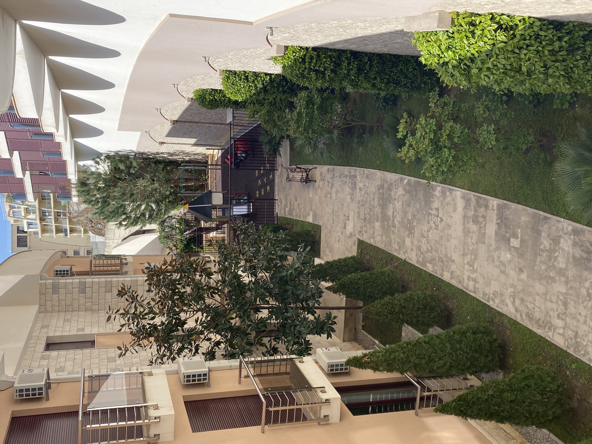The image captures a peaceful, well-maintained outdoor courtyard or common area within a residential complex or resort in Boreti, Montenegro. The scene is illuminated by bright, sunny daylight, casting sharp shadows from curved architectural overhangs, indicating late morning or early afternoon.
A winding pathway made of light-colored stone slabs meanders through a lush green landscape. The garden features meticulously kept grass, diverse shrubbery, and several trees, including a large tree with broad, dark green leaves, pine trees, and neatly shaped conical bushes lining parts of the path.
To the left of the pathway, a children's playground with a dark soft surface, a slide, and other climbing structures is visible. A dark metal bench with wooden slats sits adjacent to the playground, offering a resting spot. The surrounding buildings showcase a modern architectural style, with light-colored facades in beige and light grey stone tiles. Numerous air conditioning units are mounted on the exterior walls, with at least one clearly displaying the "TCL" brand logo. Metal railings are fitted on balconies and windows, and some windows feature brown shutters.
No people are present in the image, contributing to a serene and tranquil atmosphere. In the background, additional buildings with similar contemporary designs can be seen, suggesting an expansive complex.