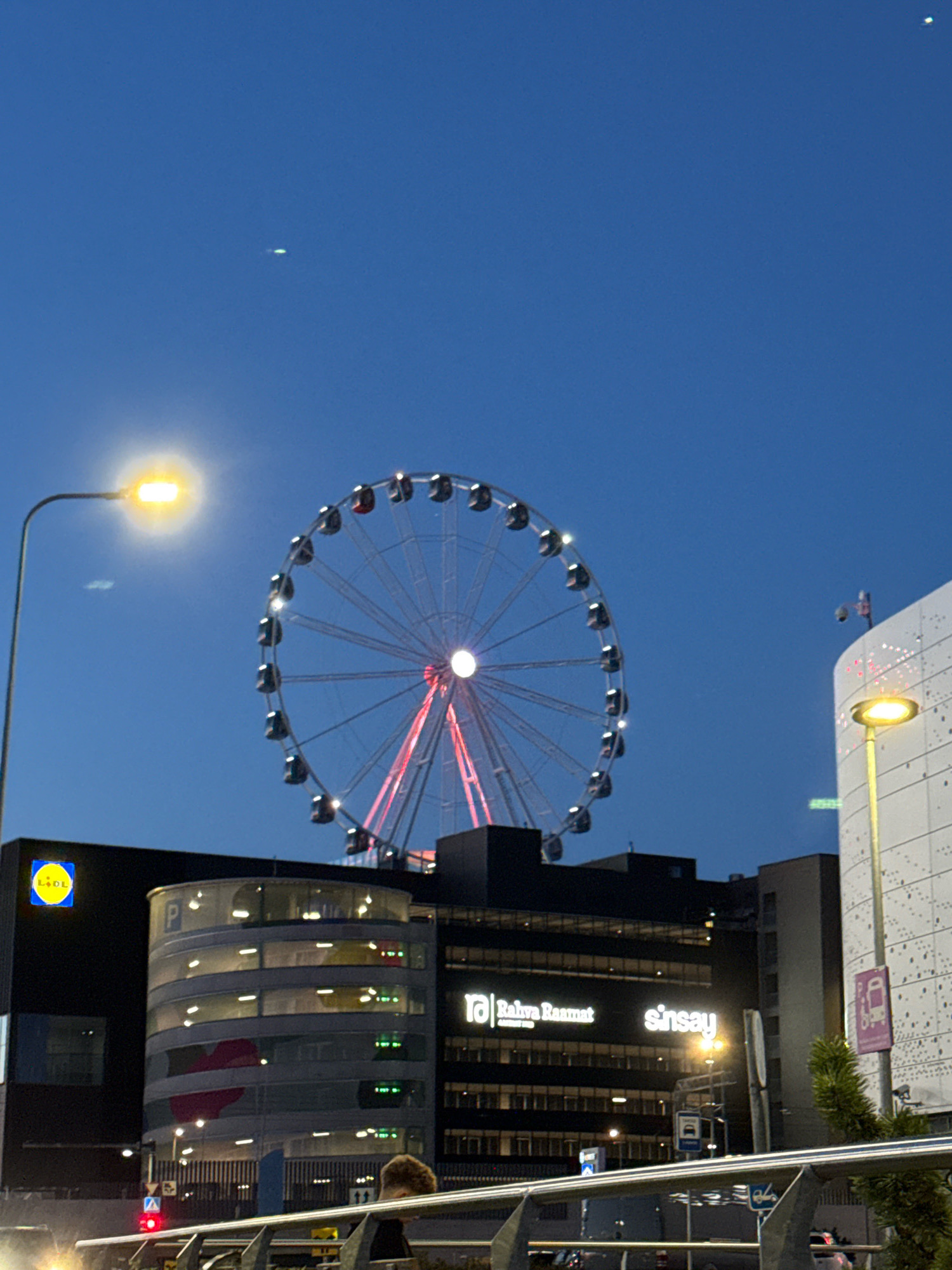 The image captures a twilight view of Tallinn, Estonia, featuring the Baltic Sea Wheel as a prominent landmark. The sky is a deep blue, indicative of the evening hour. The Ferris wheel, illuminated with red and white lights, stands tall, its structure a complex lattice of metal. Surrounding it are modern buildings, including a Lidl supermarket, recognizable by its distinctive yellow and blue logo, and a building with the Sinsay logo. The architecture is contemporary, with large windows and illuminated interiors. Streetlights cast a warm glow, and a traffic light is visible in the foreground, showing a red signal. A person is partially visible in the lower right quadrant, facing away from the camera. The overall scene depicts an urban environment at dusk, with the Ferris wheel serving as a focal point of the cityscape.