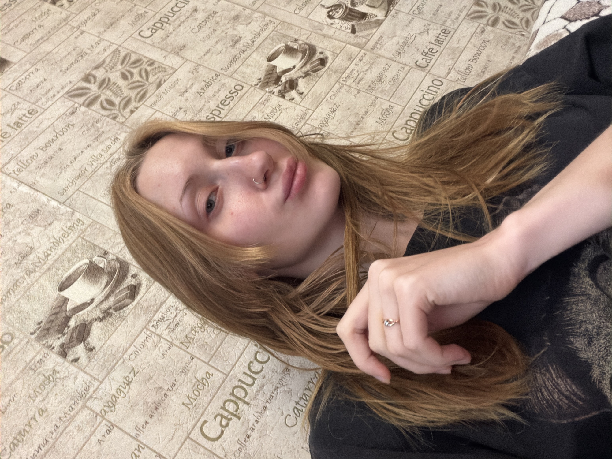 A young woman with blonde hair and a nose ring is shown in a close-up selfie. She is wearing a black t-shirt and has her hand near her face, touching her hair. She is also wearing a ring on her left ring finger. The background is wallpaper with a coffee theme, featuring images of coffee cups and text related to different coffee types. The setting is indoors, likely a residential or commercial space in Saint Petersburg, Russia. The lighting appears to be artificial.