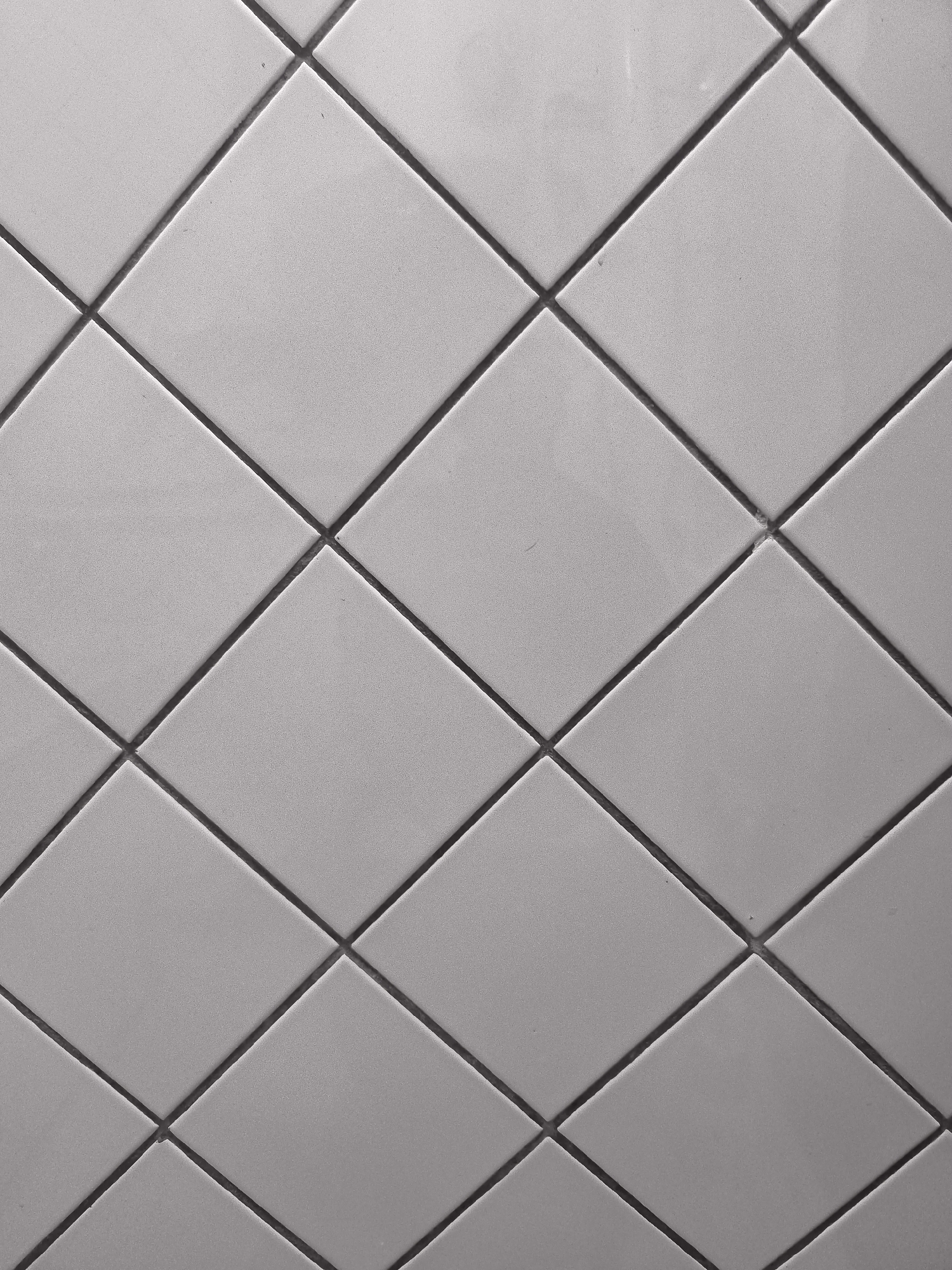 The image shows a close-up, overhead view of a tiled surface. The tiles are square and are arranged in a diamond pattern, with dark grout lines forming a grid. The tiles appear to be a neutral, light gray color and have a slightly textured surface. There are no people or any other discernible objects in the image, nor is there any indication of the specific location in Kokand, Uzbekistan. The lighting is uniform, suggesting an indoor or overcast environment.