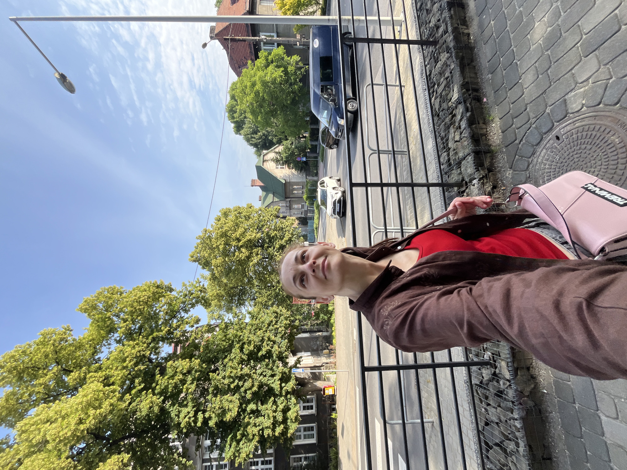 An eye-level, medium shot captures a woman with her hair pulled back, looking up and to the right with a slight smile as she holds a pink shoulder bag with the word "TRENAIL" in black lettering. She is wearing a dark brown zip-up jacket over a red tank top. The woman is standing on a cobblestone sidewalk next to a metal fence. Behind the fence, a street with traffic is visible, including a white SUV and a dark blue van. Buildings and lush green trees line the street, under a bright blue sky with scattered white clouds.