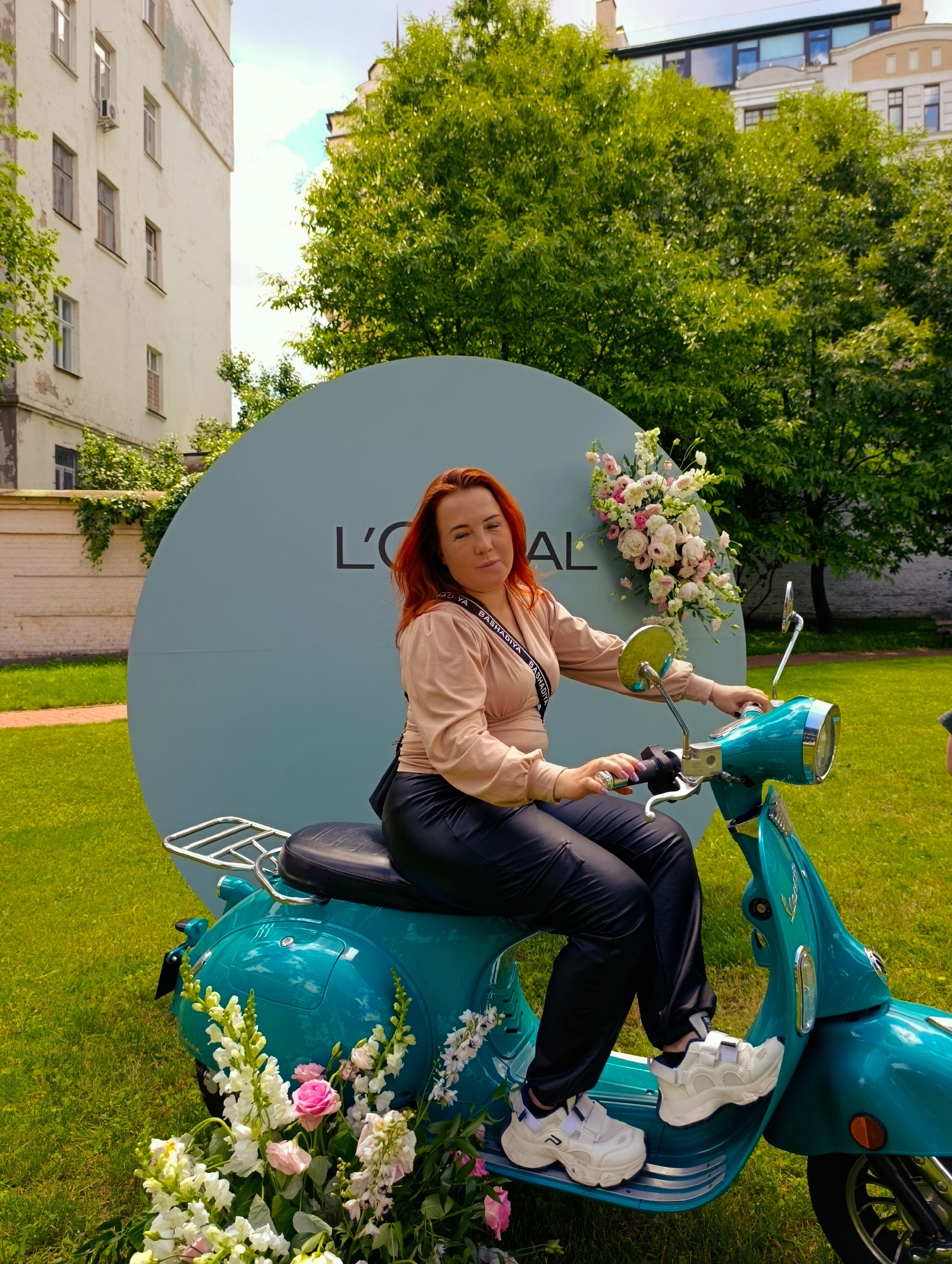 A Caucasian woman with red hair is sitting on a teal scooter in front of a large, light blue circular backdrop with the L'Oréal logo on it. She is wearing a light beige long-sleeved top, black leather pants, and white chunky sneakers. She is holding the scooter's handlebars and looking towards the camera with a slight smile. A large bouquet of pink and white flowers is arranged on the backdrop behind her, and another smaller bouquet of flowers is placed on the ground in front of the scooter. The scene is set outdoors on a grassy lawn with trees and buildings in the background, suggesting a city park or an event venue. The presence of the L'Oréal logo indicates a promotional event for the brand. The architecture of the buildings in the background, particularly the ornate details of the building on the left, could potentially help in pinpointing the exact location within Kyiv, Ukraine, but without further information or visible landmarks, a precise identification is not possible.