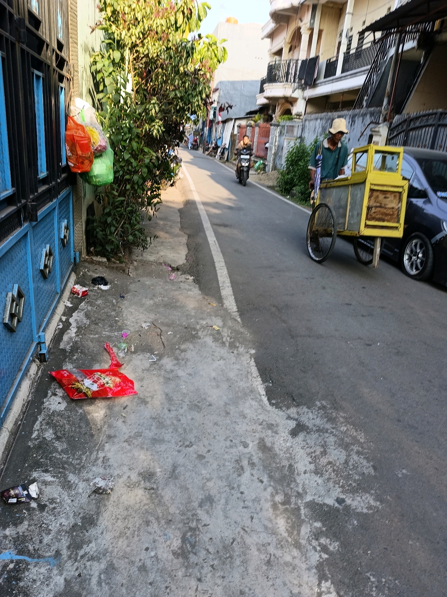 The image depicts a street scene, likely in Makassar, Indonesia, characterized by a residential or commercial environment. On the left, a blue metal fence with decorative elements runs along the edge of the sidewalk. Several plastic bags, containing what appear to be groceries or trash, are hanging from a hook on the wall near the fence. The sidewalk itself is paved with concrete and shows signs of wear and tear, with scattered litter including plastic bags and wrappers.
The street, paved with asphalt, occupies the right side of the frame. A man wearing a light-colored hat and a teal shirt is pushing a yellow cart on wheels, suggestive of a street vendor. Behind him, a dark-colored car is parked. Further down the street, a person is riding a motorcycle. The background reveals multi-story buildings with balconies and railings, typical of urban architecture. Trees and foliage are visible along the street and between buildings, suggesting a mix of urban and green spaces. The lighting indicates it is daytime, with sunlight casting shadows and illuminating the scene. The overall impression is of a moderately busy urban street with elements of daily life, commerce, and the presence of litter.