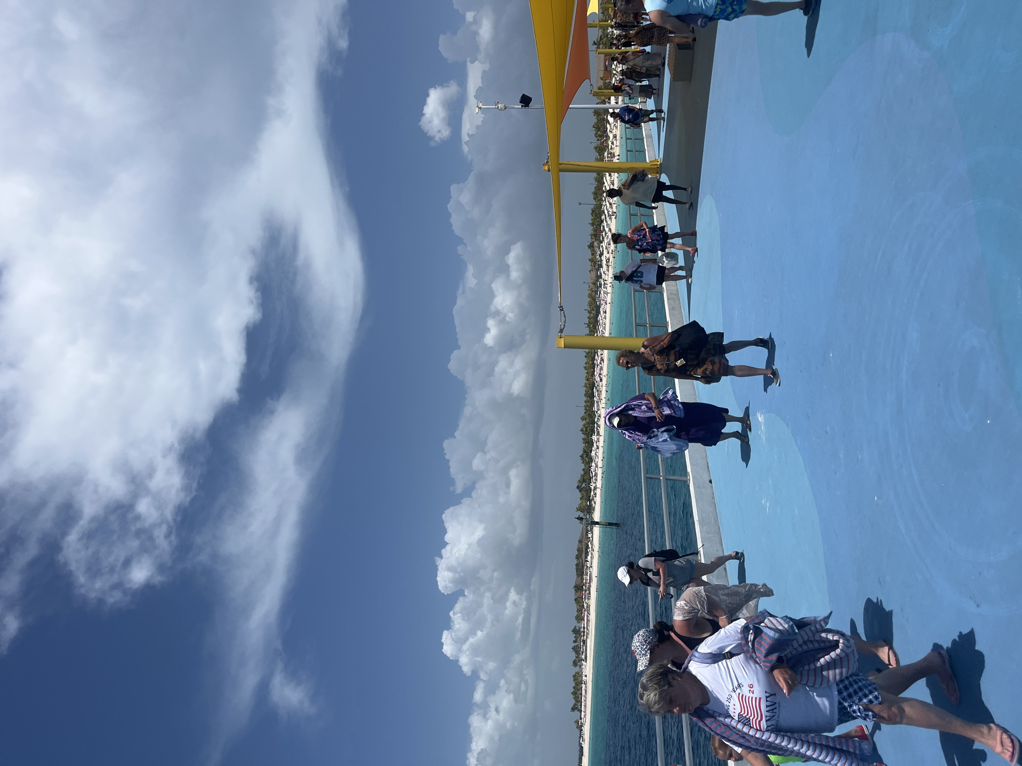 The image shows a bright, sunny day on a pier in the City of Freeport, Bahamas. A group of people are walking along a bright blue, wavy-patterned surface. In the background, the turquoise ocean meets a white sandy beach lined with palm trees and resort buildings. Several small boats and umbrellas are visible on the beach. The sky is a vibrant blue with scattered white, fluffy clouds. The people on the pier are dressed in casual, summery clothing, with some carrying bags or towels. The pier itself has a metal railing along the side, and part of the pier is covered by a bright yellow awning. The overall scene depicts a lively tropical vacation destination.