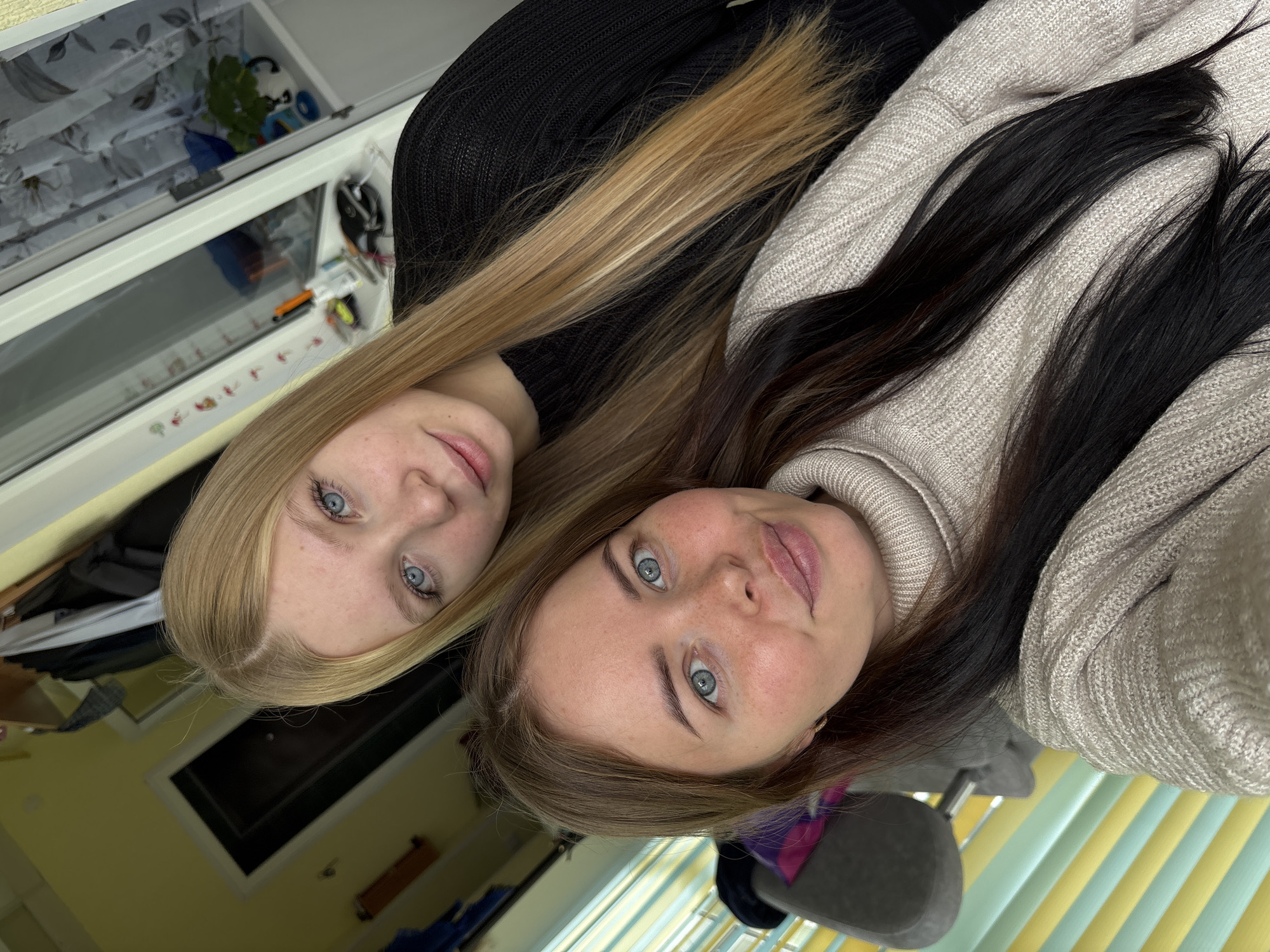 This image, potentially taken in Chaikovsky, Russia, features two young women posing for a portrait indoors. They appear to be lying down, looking upwards directly at the camera, which suggests a selfie. Both subjects have calm, neutral expressions with a slight hint of a smile.
The woman on the left has long, straight blonde hair and bright blue eyes. She is wearing a black knitted sweater. The woman on the right has long, dark brown hair and blue eyes, with light freckles visible across her nose and cheeks. She is dressed in a light beige or cream-colored knitted turtleneck sweater.
The background reveals a casual indoor setting. To the upper left, a white cabinet with reflective or glass doors contains various items, including what looks like a white plush toy, some green foliage, and other indistinct objects. On a lower shelf, small colorful items are visible, but no clear text can be deciphered. To the lower right, striped window blinds or curtains in alternating light yellow/beige and light blue/green hues indicate natural daylight entering the room. A grey piece of furniture, possibly a chair or part of a bed, is also visible. The women's warm sweaters suggest an indoor setting that may be cool. No discernible text is present in the image.
