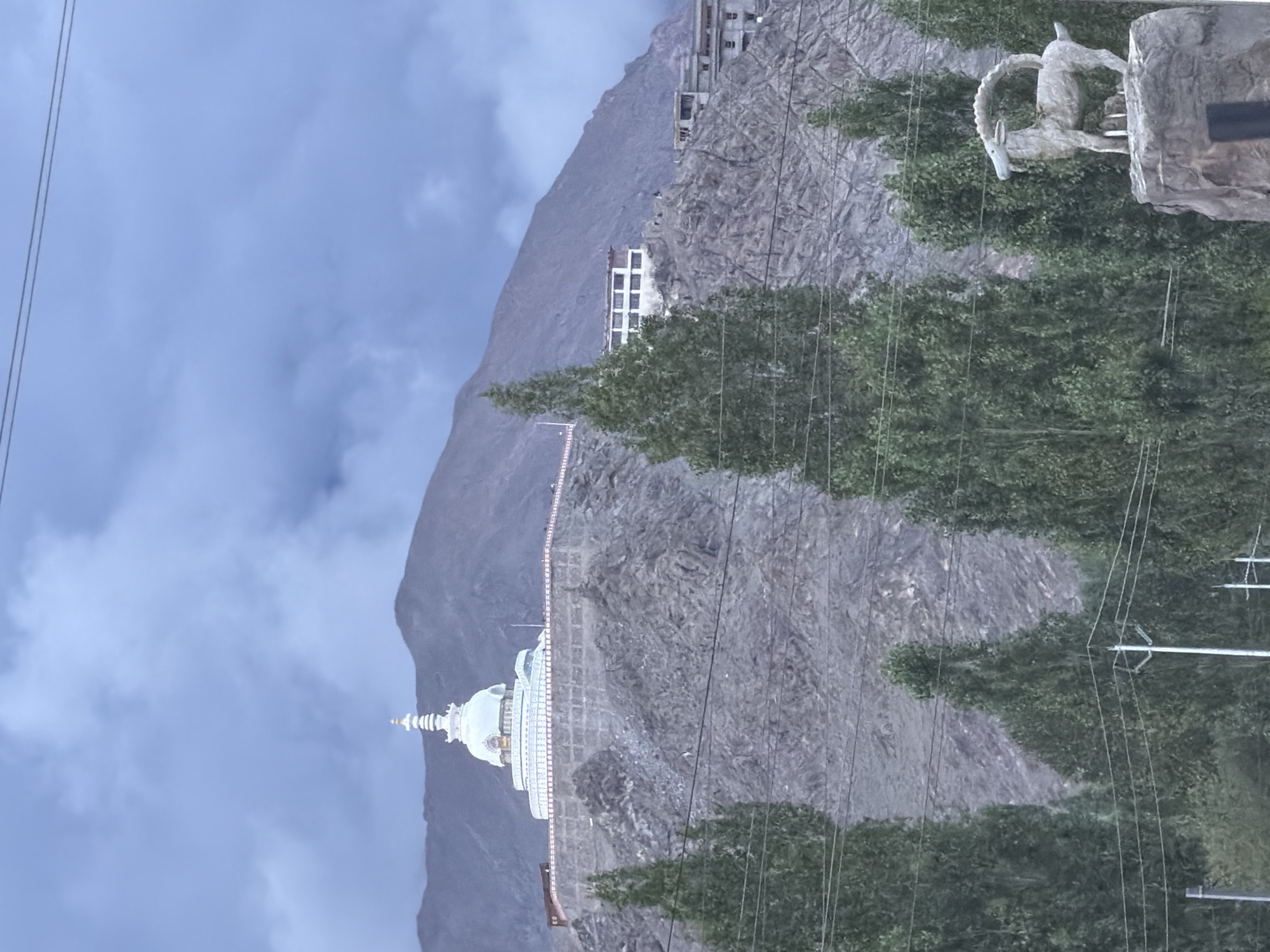 The image shows the Shanti Stupa in Leh, India. The stupa is a white Buddhist monument located on a hilltop, overlooking the city. It is surrounded by a rocky, mountainous landscape, with trees in the foreground. The sky is cloudy and overcast. In the lower right corner, there is a statue of a goat. The scene appears to be a tourist attraction.