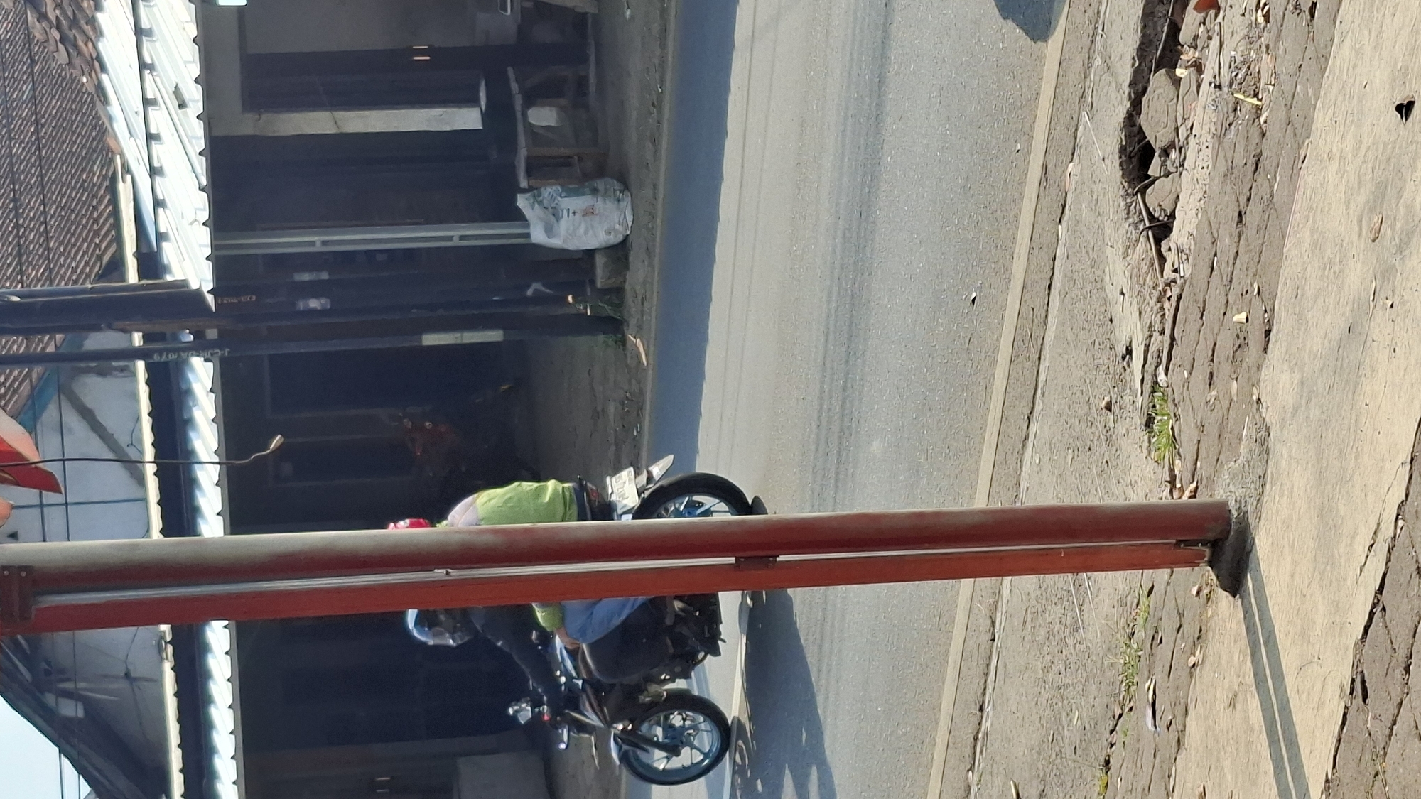 A motorcyclist wearing a helmet and a light green jacket rides a dark motorcycle with blue accents on the wheels. They are on a paved road. In the background, there is a building with a tiled roof and a white metal awning. To the right of the building, there is a white bag of what appears to be cement or similar material. A red pole stands prominently in the foreground, slightly off-center to the left. The ground next to the road consists of broken pavement and some sparse vegetation. The scene is well-lit by sunlight, casting shadows. This image was taken on Jalan Raya Cilaku in Cilaku, Indonesia.