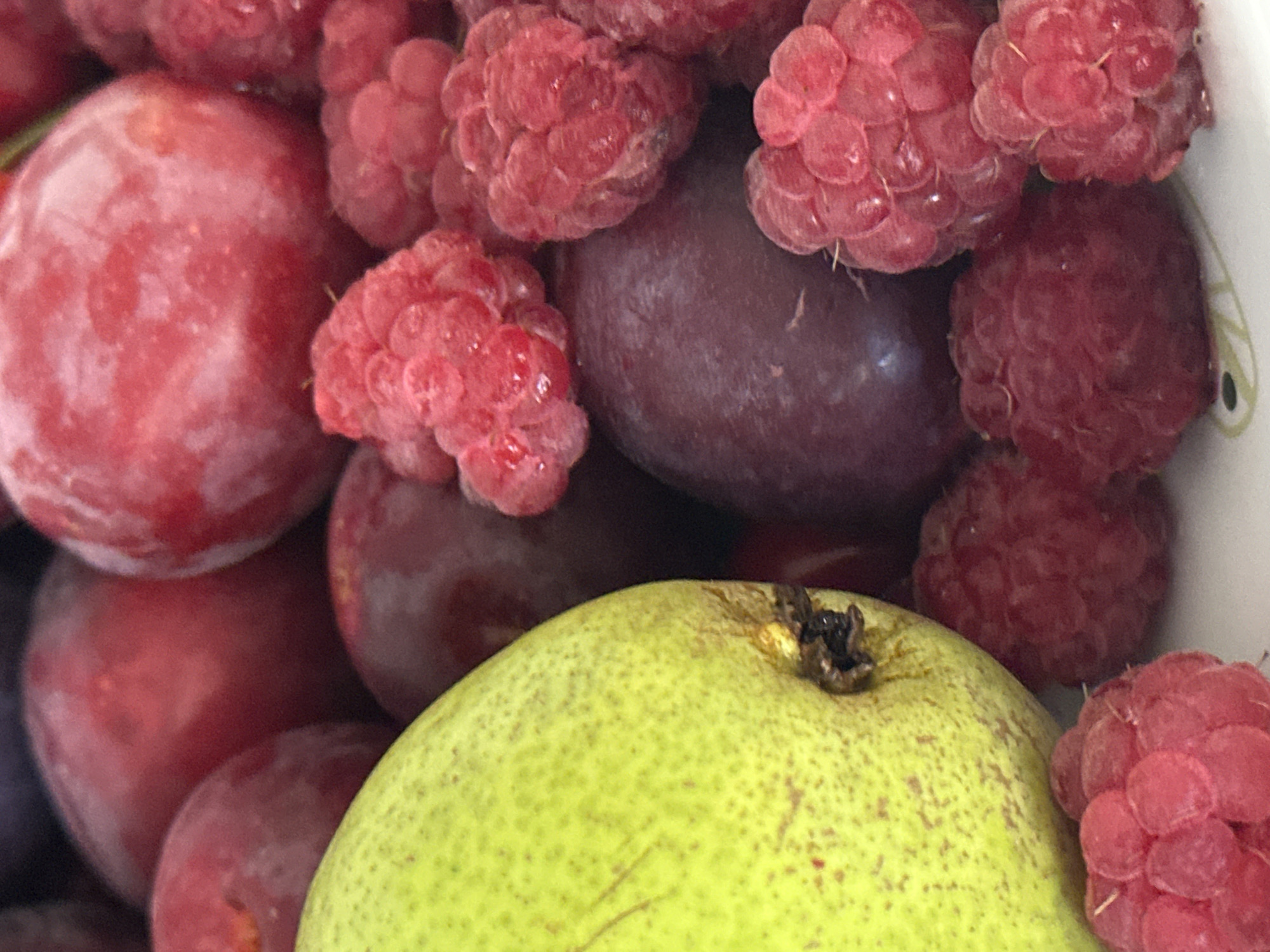 A close-up, overhead shot showcases a vibrant assortment of fresh fruits, including a bright green pear, deep red plums, and plump raspberries. The fruits are artfully arranged, with the pear occupying the left side of the frame and the plums and raspberries filling the rest of the image. The soft lighting highlights the natural textures and rich colors of the fruits, creating a visually appealing and appetizing scene. The fruits are likely at a market stall in Nevinnomyssk, Russia, or a local grocery store.
