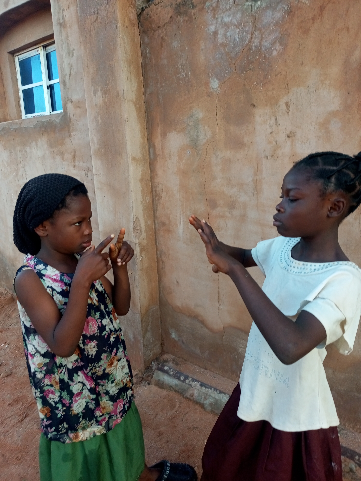 Two young girls are standing outside a building with a rough, textured, tan-colored wall in Tempure, Nigeria. The girl on the left, wearing a black beanie and a floral-patterned sleeveless dress over a green skirt, is gesturing with both hands, her index fingers extended and pointing. She has her head tilted and is looking towards the girl on the right. The girl on the right, with her hair in braids, is wearing a white top with a decorative neckline and a dark red skirt. She is holding her hands up in front of her, palms facing outward, and also appears to be gesturing. In the background, to the left, is a small window with white framing and bright blue panes. The ground is covered in reddish-brown dirt.