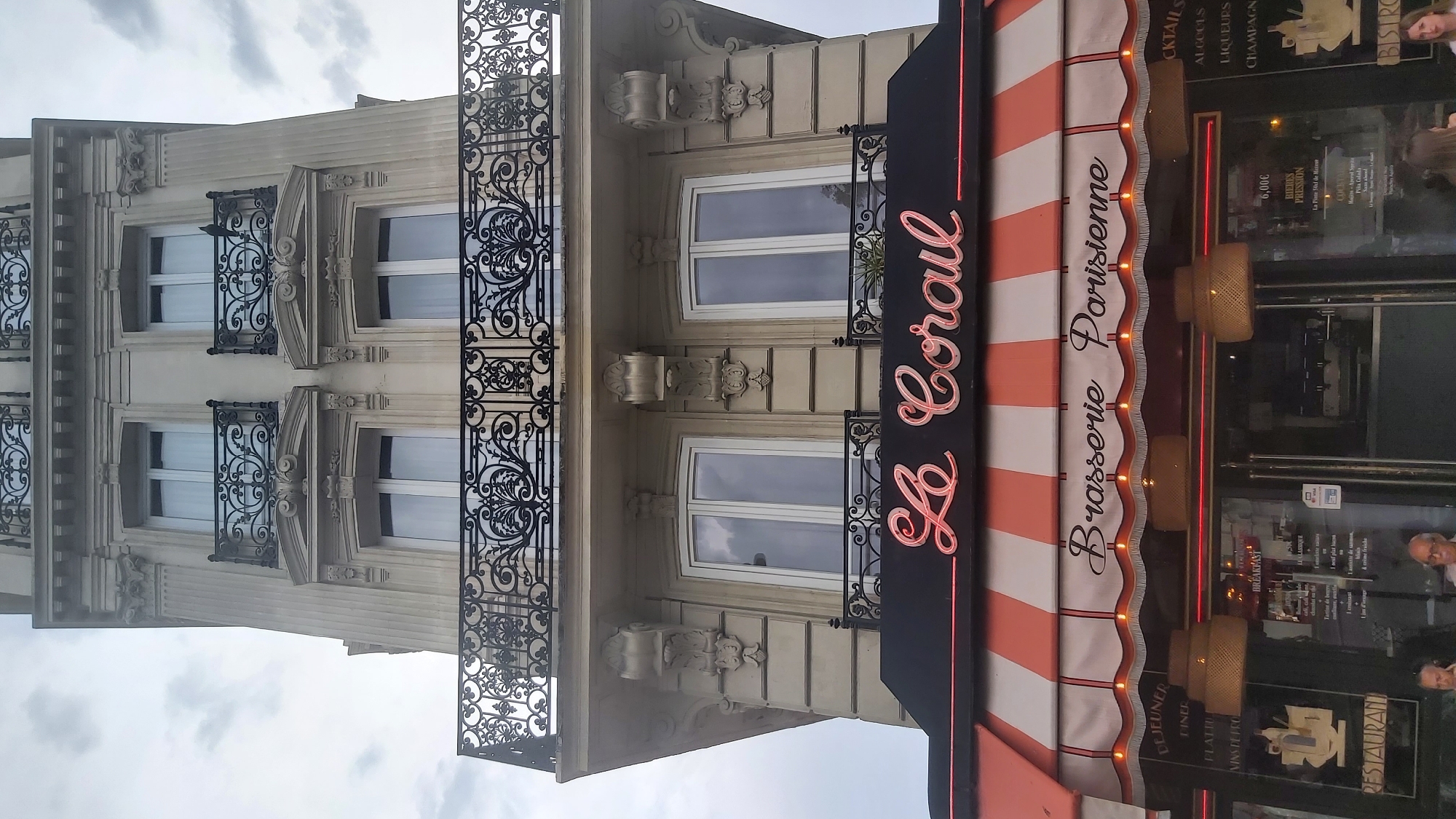 The image captures a detailed low-angle shot of a Parisian building facade, focusing on the upper floors and the storefront below. The building exhibits classic Haussmannian architecture with stone cladding, ornate balconies with wrought-iron railings, and large windows. The prominent storefront displays a neon sign that reads "Le Corail," with "Brasserie Parisienne" written below it in script. An orange and white striped awning stretches across the storefront, decorated with small lights. The windows of the brasserie reveal the interior, with patrons visible inside. The sky above is overcast, suggesting a cloudy day in Paris.