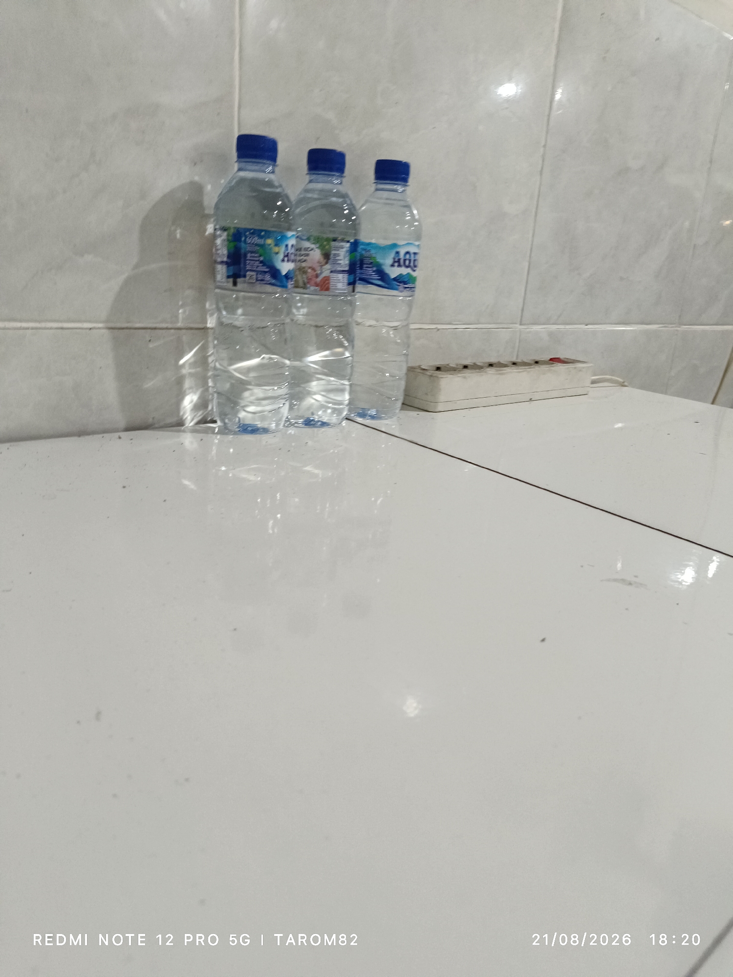 In the city of Sidodadi, Indonesia, a photograph captures a tiled wall with three bottles of Aqua water lined up on a white surface. To the right of the water bottles, a white electrical power strip with multiple sockets is visible. The lighting in the scene suggests it is indoors. The bottom of the image displays text indicating "REDMI NOTE 12 PRO 5G I TAROM82" and "21/08/2026 18:20".