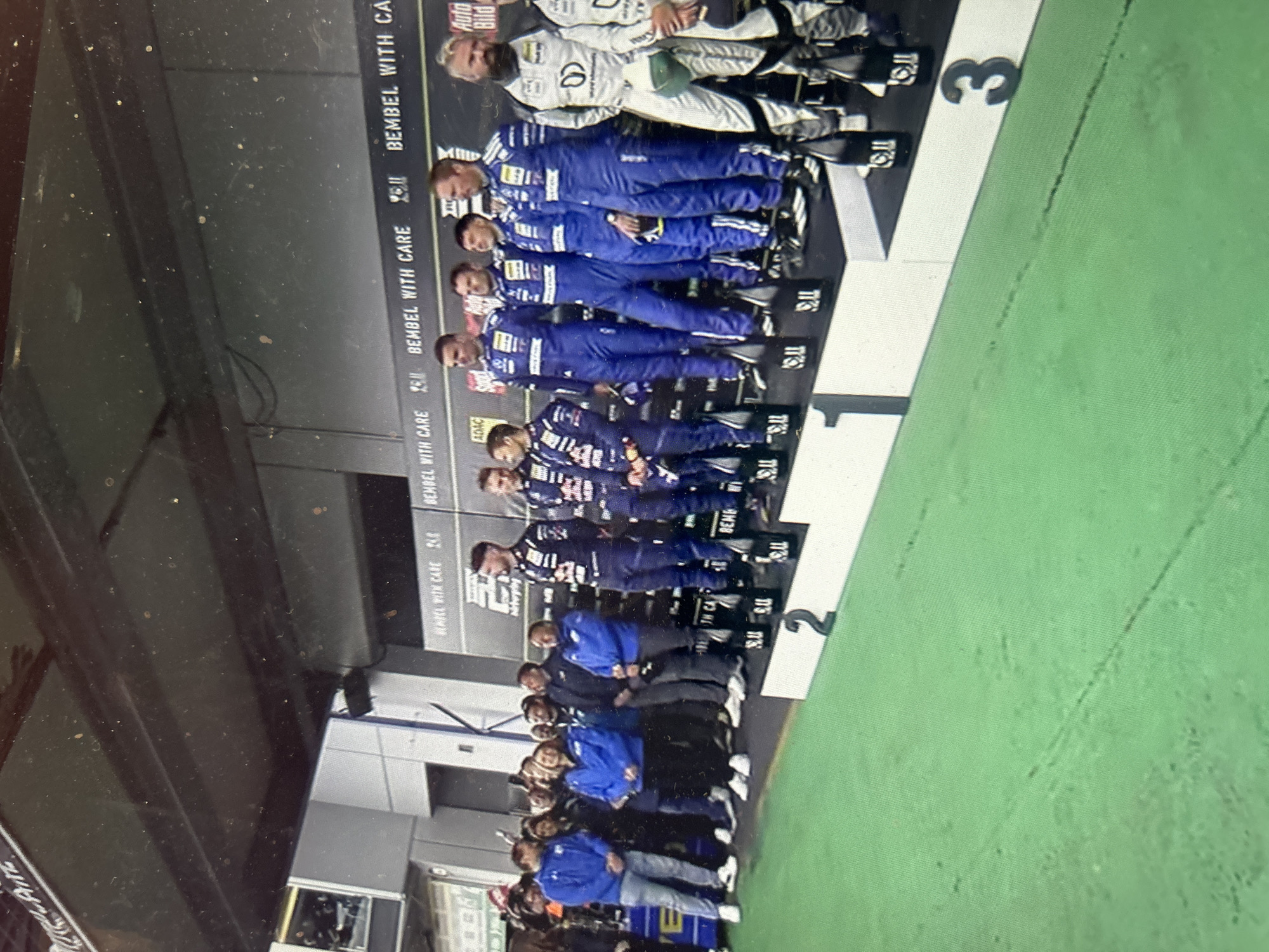 The image shows people standing on a podium. The podium has labels "1," "2," and "3." The people on the podium are dressed in racing suits and jackets. Some of the racing suits are white with black accents, while others are blue with white accents. Above the podium is a sign that reads "BEMBEL WITH CARE." The environment appears to be indoors, possibly a garage or event space.