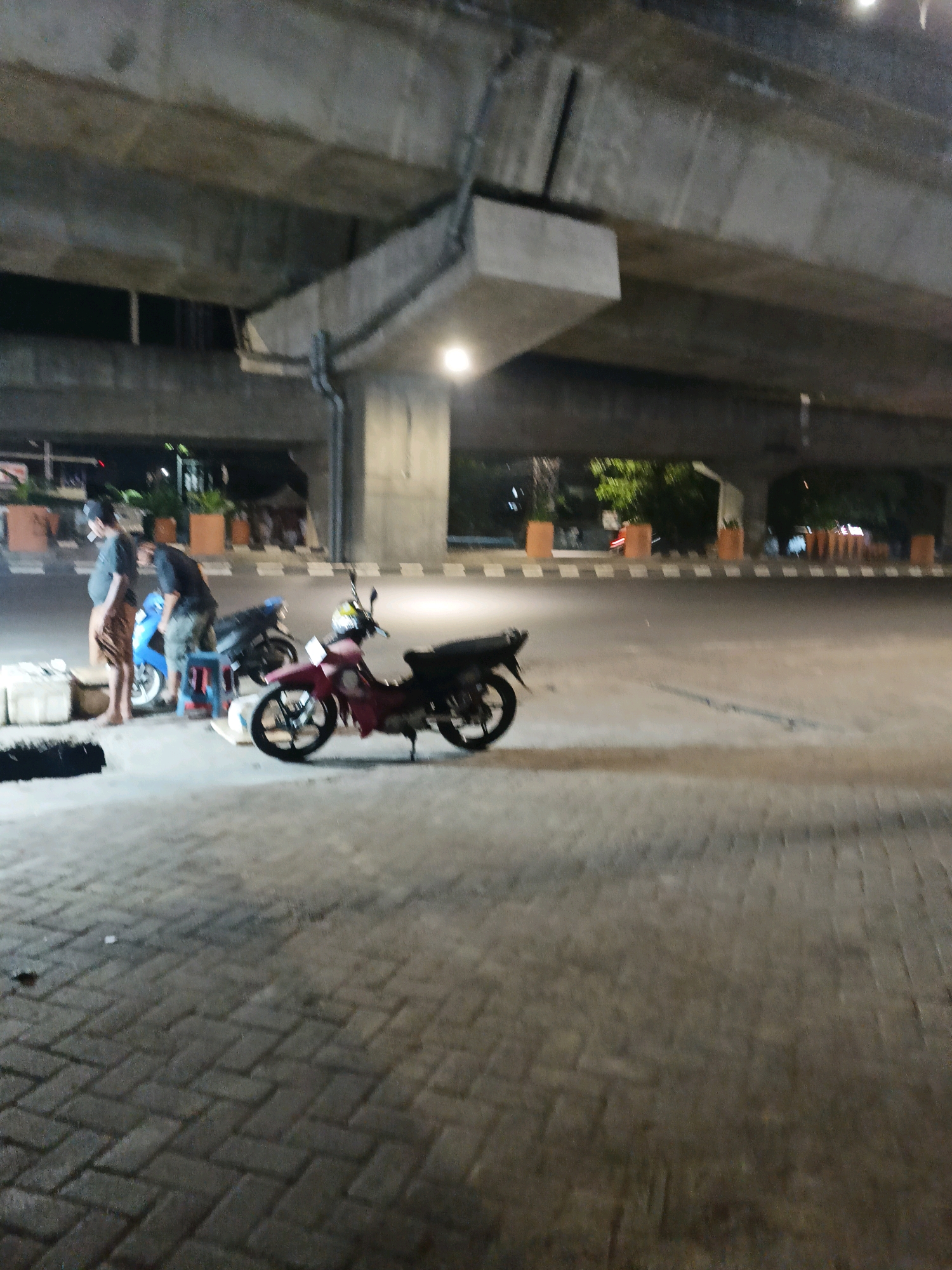 The image depicts an outdoor scene at night, likely in Makassar, Indonesia, underneath a concrete overpass. In the foreground, a paved area with interlocking bricks is visible. To the left, two individuals are standing near a light blue motorcycle and a dark motorcycle. One individual, wearing a dark shirt and a sarong, is facing left. The other individual, in a dark shirt and pants, is looking towards the motorcycles. A red motorcycle is parked in the center of the image, with its kickstand down. The background shows the underside of the overpass, with support structures and what appears to be lighting. Beyond the overpass, trees and some structures are faintly visible.