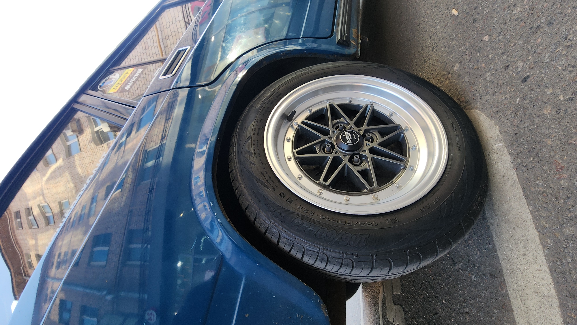 An eye-level, close-up, low-angle exterior shot captures the rear wheel and side panel of a dark blue, older model sedan. The car is parked on a paved surface marked with a light-colored stripe, likely a parking space or crosswalk, indicating an urban setting. In the background, a brick building with multiple windows is visible, its reflection distorted in the car's polished surface. The building appears to be in a state of disrepair, with some windows boarded up and the brickwork showing signs of age. A sticker with Cyrillic text, including the word "АДАПТЕРОВ," is affixed to the rear window. The wheel itself is a multi-spoke, aftermarket design with a silver lip and black spokes, featuring the "Equip" logo on the center cap. The tire is a black radial, with visible text including "185/60R14 82H." The overall scene suggests a potentially older, urban environment, possibly in Moscow, Russia, given the Cyrillic script. No people are present in the frame.