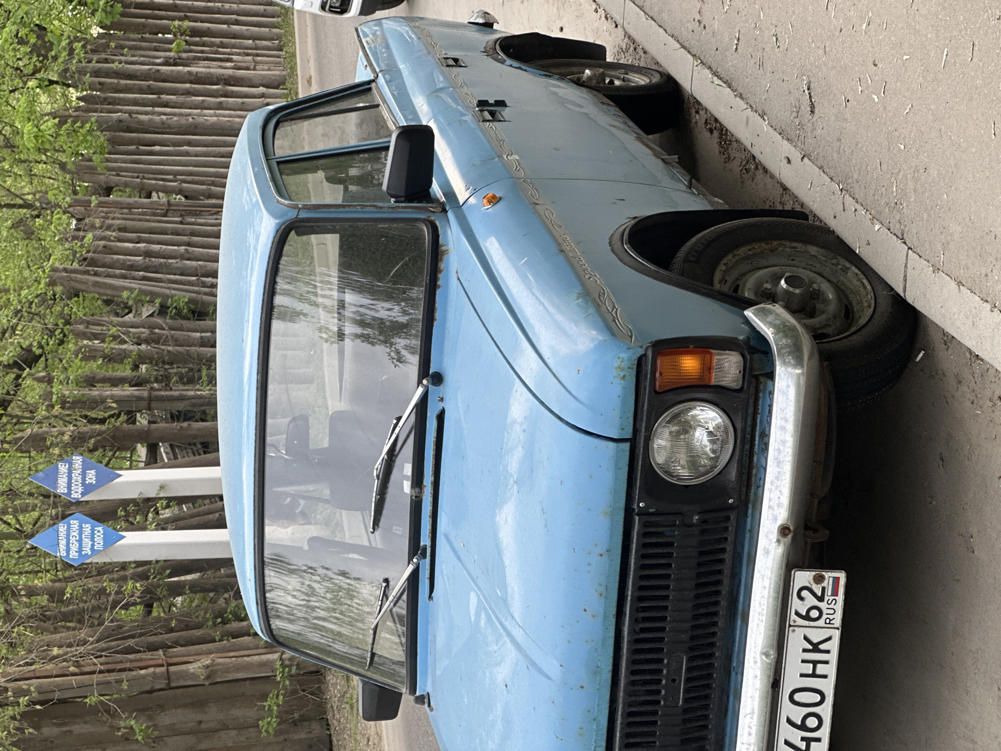 The image depicts a blue, old, possibly Soviet-era car parked on the side of a street. The car has visible rust and wear. The license plate reads "460 HK 62 RUS," indicating it is registered in Russia. The scene includes trees in the background and signs in Russian. Given the license plate and signage, the location is likely in Russia, and judging from the style of the license plate, is likely in or near Moscow. The environment is urban, evidenced by the presence of the street and the parked car. There are no people visible in the frame.