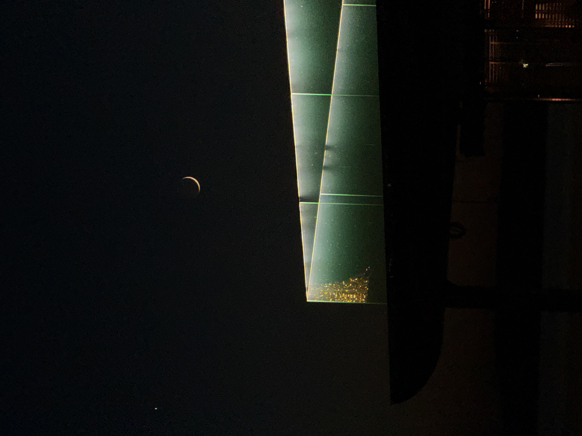 The image depicts a night scene in Votkinsk, Russia. A crescent moon is visible in the dark sky, and a bright dot, likely a star or planet, appears below it. To the right of the sky is an illuminated building, with stacked, partially lit green rectangular structures and some darker, presumably shadowed areas. The specific location within Votkinsk is not discernible from the image.