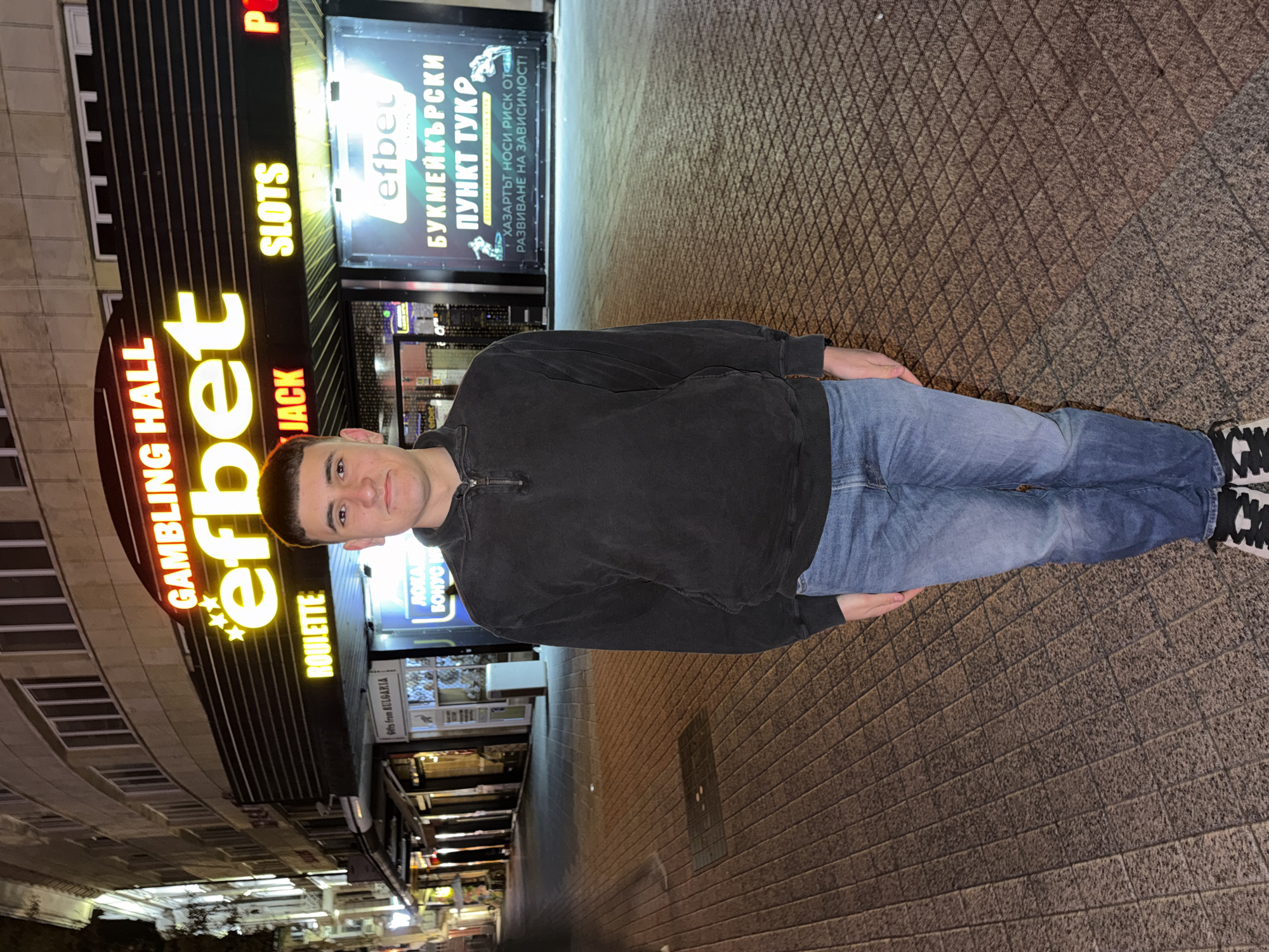 A person is standing on a textured pavement in front of a building with "GAMBLING HALL efbet" in illuminated yellow letters above a section listing various games. A sign for "efbet" is displayed in Bulgarian, accompanied by a cautionary statement about the risks of gambling addiction. The building is located in Plovdiv, Bulgaria, based on the "Gifts from BULGARIA" sign and the Bulgarian text on the gambling sign. The person is wearing a dark sweatshirt and blue jeans with black and white shoes.