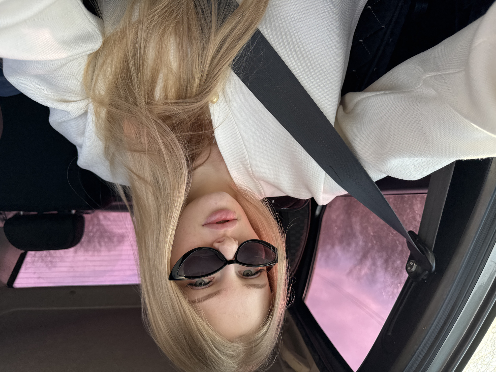 A young, fair-skinned female with long, straight, blonde hair and dark, cat-eye sunglasses is depicted seated in the front passenger position of a vehicle in Кондратово, Russia. She wears a white, textured, long-sleeved garment, with a black seatbelt fastened across her left shoulder and torso. Her gaze is directed towards the camera, with slightly parted lips. The vehicle's interior exhibits dark gray trim, a visible seatbelt anchor on the B-pillar, and a black rear headrest. Through the vehicle's windows, a prominent pinkish-purple tint or lighting effect is observed, revealing indistinct outlines of external vegetation against a bright sky.