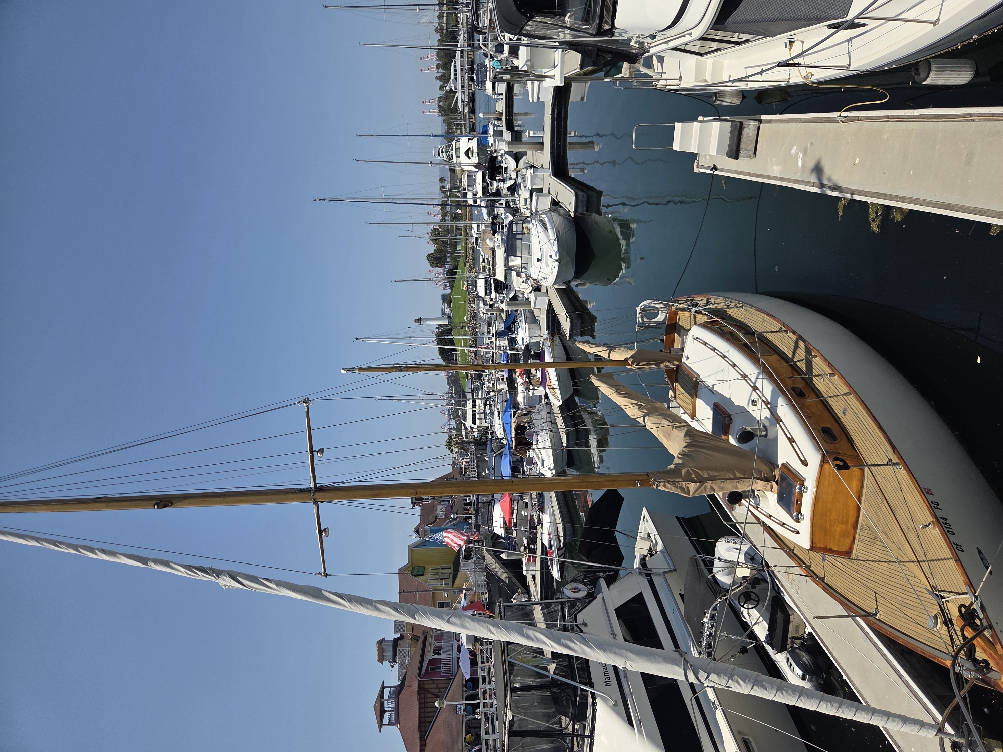 This is a high-angle, wide shot of a marina on a clear, sunny day. The foreground features a classic wooden sailboat with white sails furled. The boat is moored in a waterway alongside other vessels. In the background, a collection of buildings with colorful facades lines the waterfront, reminiscent of a commercial or entertainment district. Beyond the immediate harbor, a vast expanse of water is dotted with numerous sailboats and motorboats, their masts reaching towards the clear blue sky. In the distance, a lighthouse and industrial cranes are visible, suggesting the proximity of a port or industrial area. The scene is devoid of any people. The water in the marina is calm and reflects the sky and surrounding boats. The overall impression is one of a tranquil, sun-drenched day at a busy harbor.