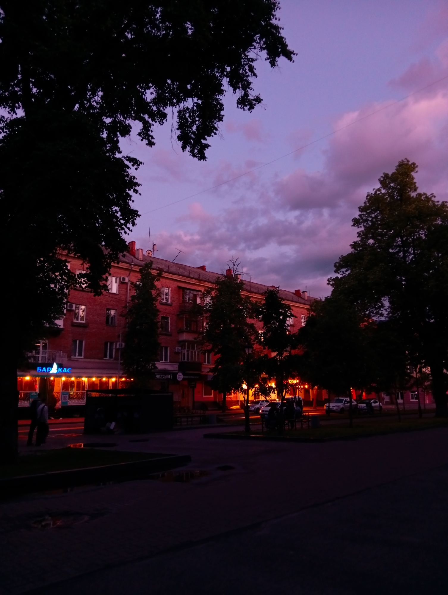 The image captures an urban street scene during twilight or early evening, characterized by a striking purple and pink sky with scattered clouds. The foreground is dark, featuring a paved area with subtle reflections, possibly from moisture. Large, dark trees frame the top and sides of the composition, their dense foliage silhouetted against the colorful sky and illuminated elements below.
A multi-story brick building stands prominently on the left side of the street, with several windows and balconies visible on its upper floors. The ground floor of this building is brightly lit with warm orange and yellow lights, indicative of businesses such as shops or cafes. A clear blue sign on the building displays Cyrillic text, reading "БАР КАС" (BAR KAS). People are visible on the sidewalk in front of these establishments; some are walking, and others appear to be seated at outdoor tables, engaging in evening social activities.
The street itself runs parallel to the building, with several cars parked along its right side further down. More trees line the street, interspersed with the parked vehicles. The overall lighting creates a strong contrast between the vibrant sky and the warmly lit commercial fronts against the encroaching darkness. Utility lines are faintly discernible against the twilight sky. The scene conveys a peaceful yet active urban evening, with the warm glow from the establishments inviting a sense of community and the dramatic sky adding a touch of natural beauty. The Cyrillic text on the signage aligns with the location context of Poltava, Ukraine.