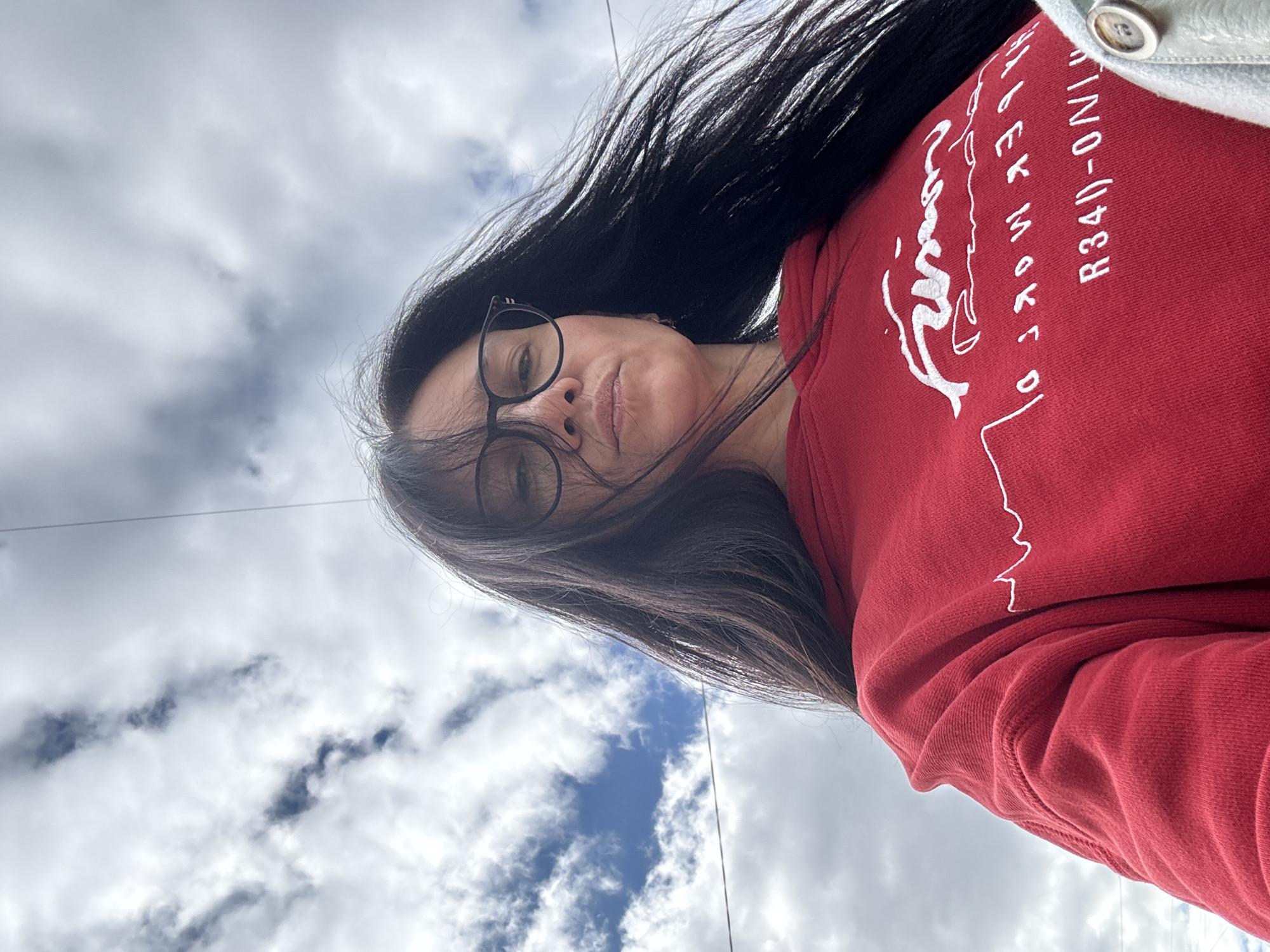 A woman with long dark hair and glasses is wearing a red hooded sweatshirt and a light-colored jacket. She appears to be looking down at the camera, which is positioned at a low angle. The background shows a cloudy sky with patches of blue visible between the clouds. Based on available information, the location appears to be in the city of Chaikovsky, Russia, as indicated by the text "R340-ONI" on the sweatshirt, which could be related to a local business or brand.