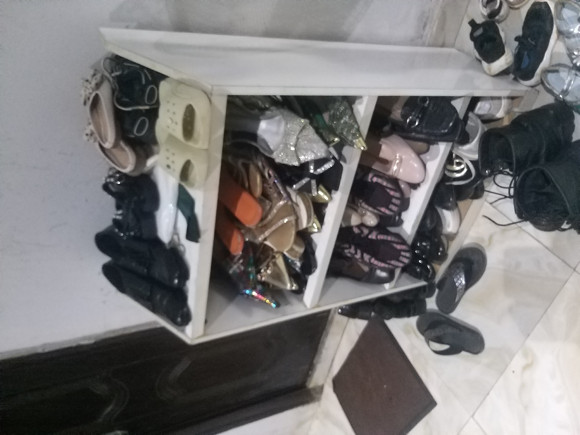 The image depicts a white, multi-tiered shoe rack filled with various footwear. The top shelf holds a pair of black formal shoes and a pair of black open-toed sandals. The second shelf displays a mix of sandals and heels, including some with embellishments and others in solid colors. The third shelf contains more sandals and shoes, some with decorative elements. The bottom shelf of the rack holds a pair of black formal shoes, a pair of black slip-on shoes, and a pair of pink ballet flats. On the floor next to the shoe rack, there are two black flip-flops. Further to the right, a collection of sneakers and athletic shoes are arranged. The background shows a portion of a dark-colored door and a light-colored wall. The scene appears to be indoors, on a tiled floor. The image is likely from Kano, Nigeria, given the prompt, but there are no visual cues within the image itself to confirm this specific location.
